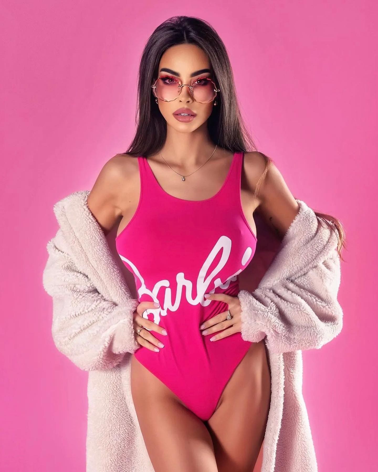 Standing against a pink backdrop, she wears a pink swimsuit with white lettering, a fluffy coat, and pink glasses.