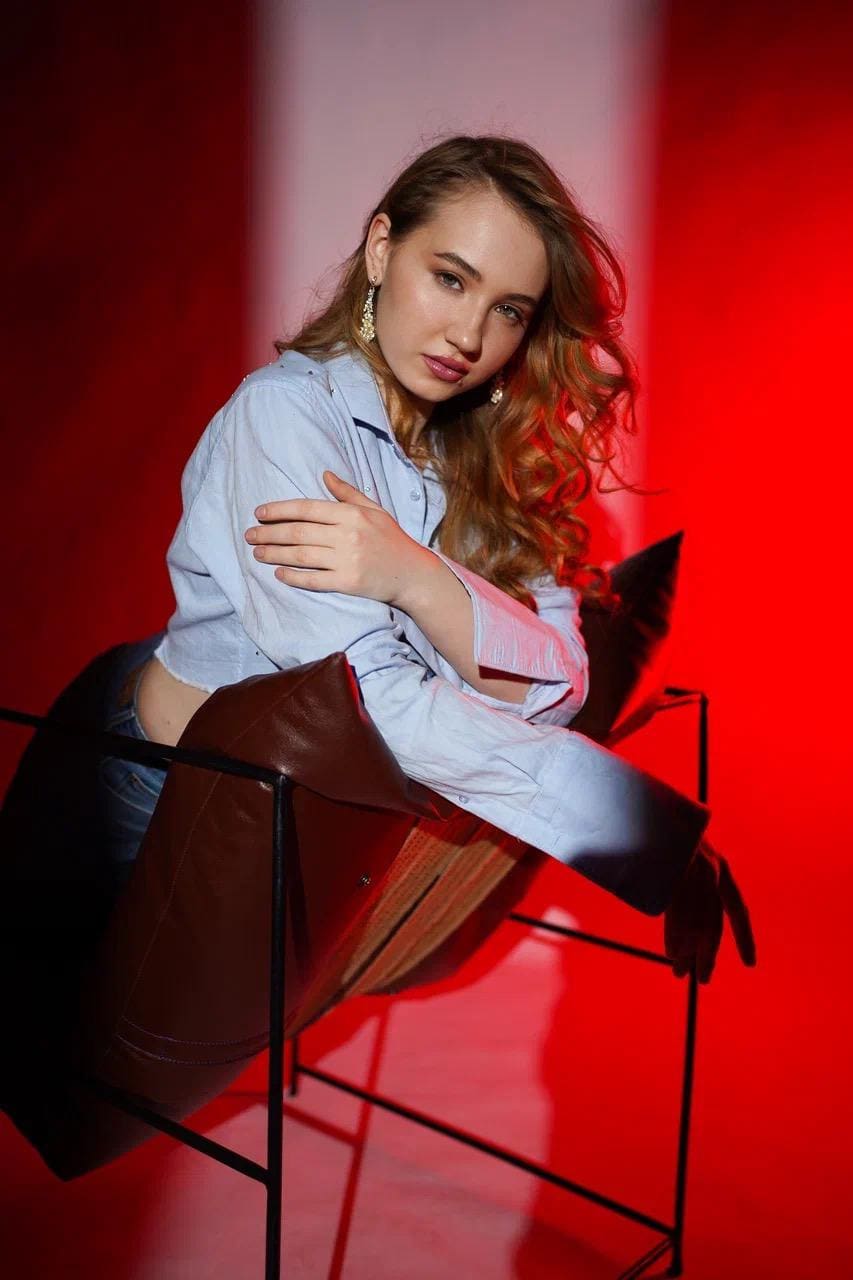 A woman in a light blue shirt and jeans sits on a black chair against a red background, arms crossed on a cushion.