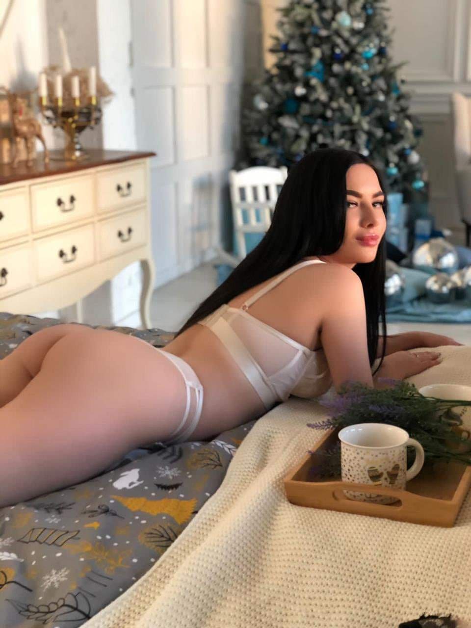 A woman with long dark hair lies on her stomach on a bed wearing a white bra and underwear, with a Christmas tree and dresser in the background.