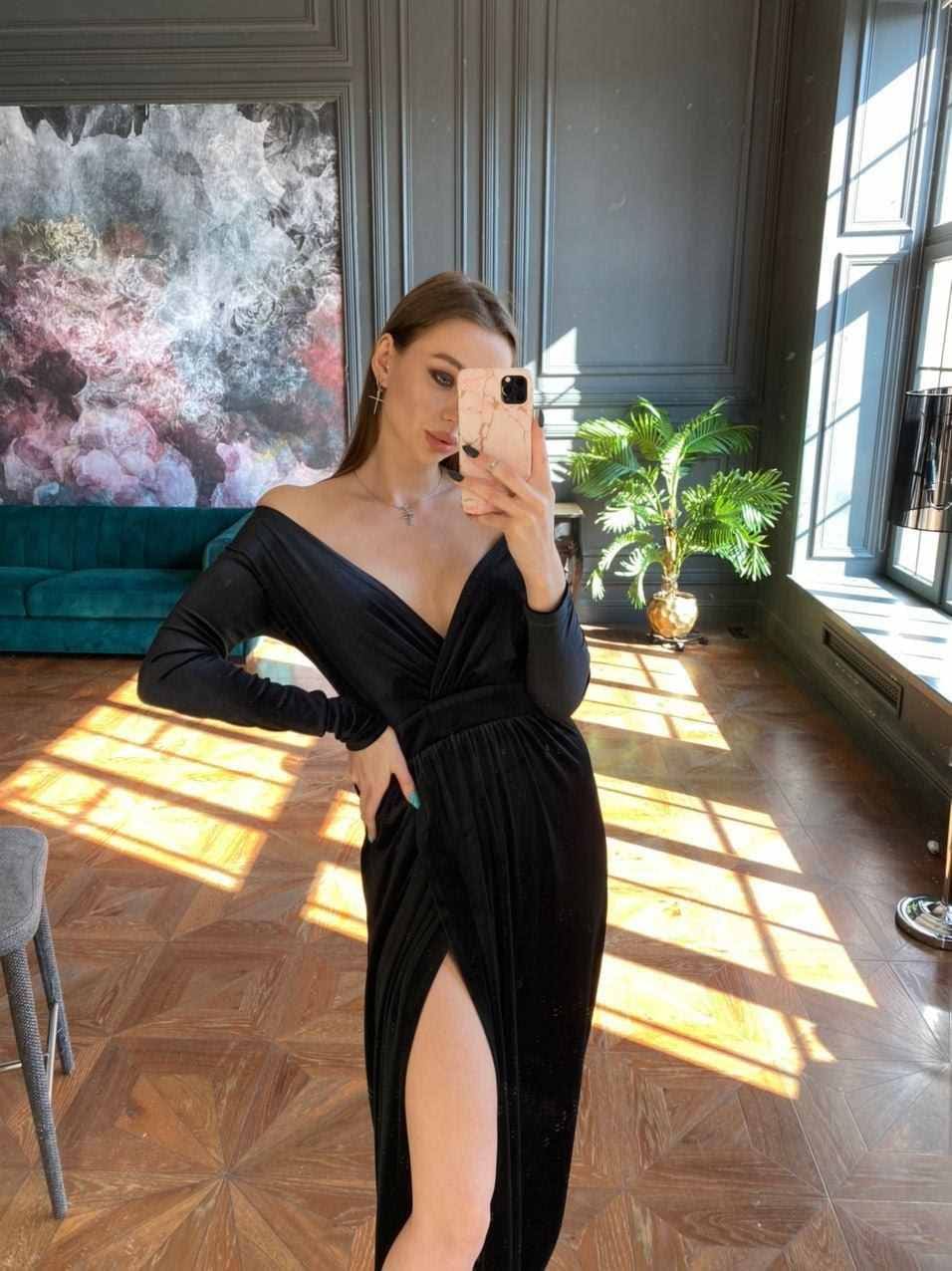Woman in a black velvet gown with a slit takes a mirror selfie in a sunlit room with dark walls and a palm.