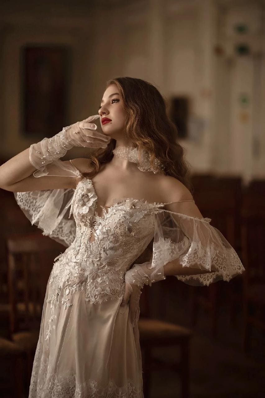 A woman stands in a dim room wearing a white lace gown, sheer gloves, and a choker, with one hand raised near her face.
