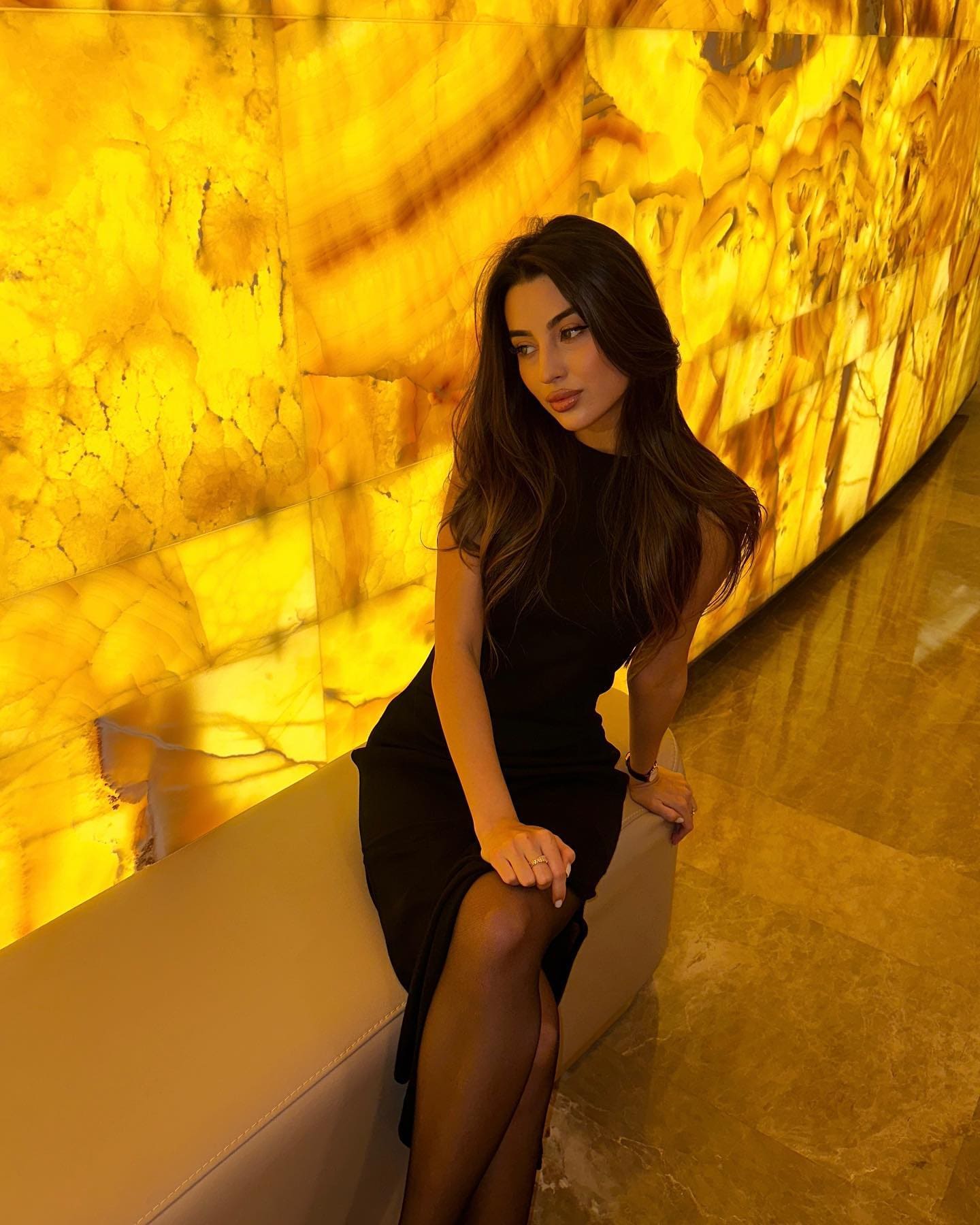 A woman sits on a white bench against a glowing yellow stone wall, wearing a sleeveless black dress with a thigh-high slit and sheer tights.
