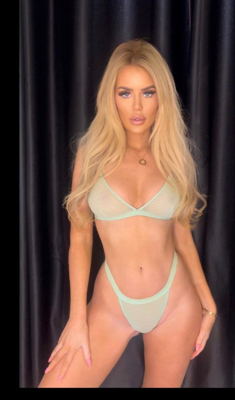 A woman in a light green two-piece bikini faces the camera against a dark curtain, resting one hand on her thigh.