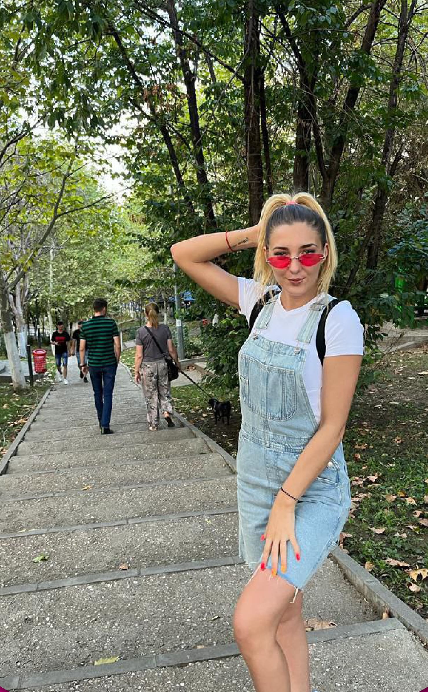 A blonde woman in denim overalls and sunglasses stands on a park path with pedestrians and a dog among trees behind her.