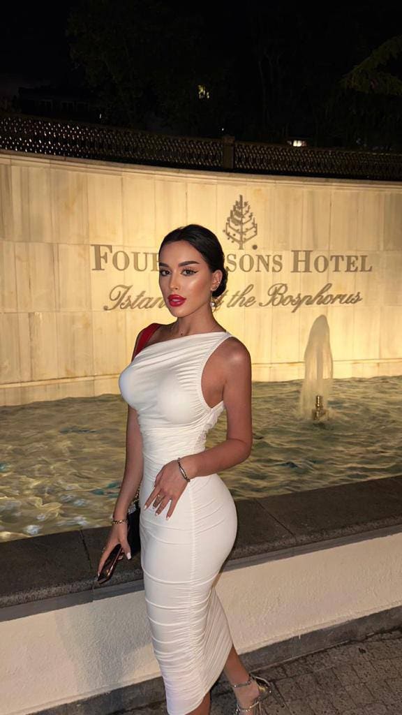 A woman stands by a fountain at night in a white one-shoulder dress with red strap detail, holding a phone and wearing heels.