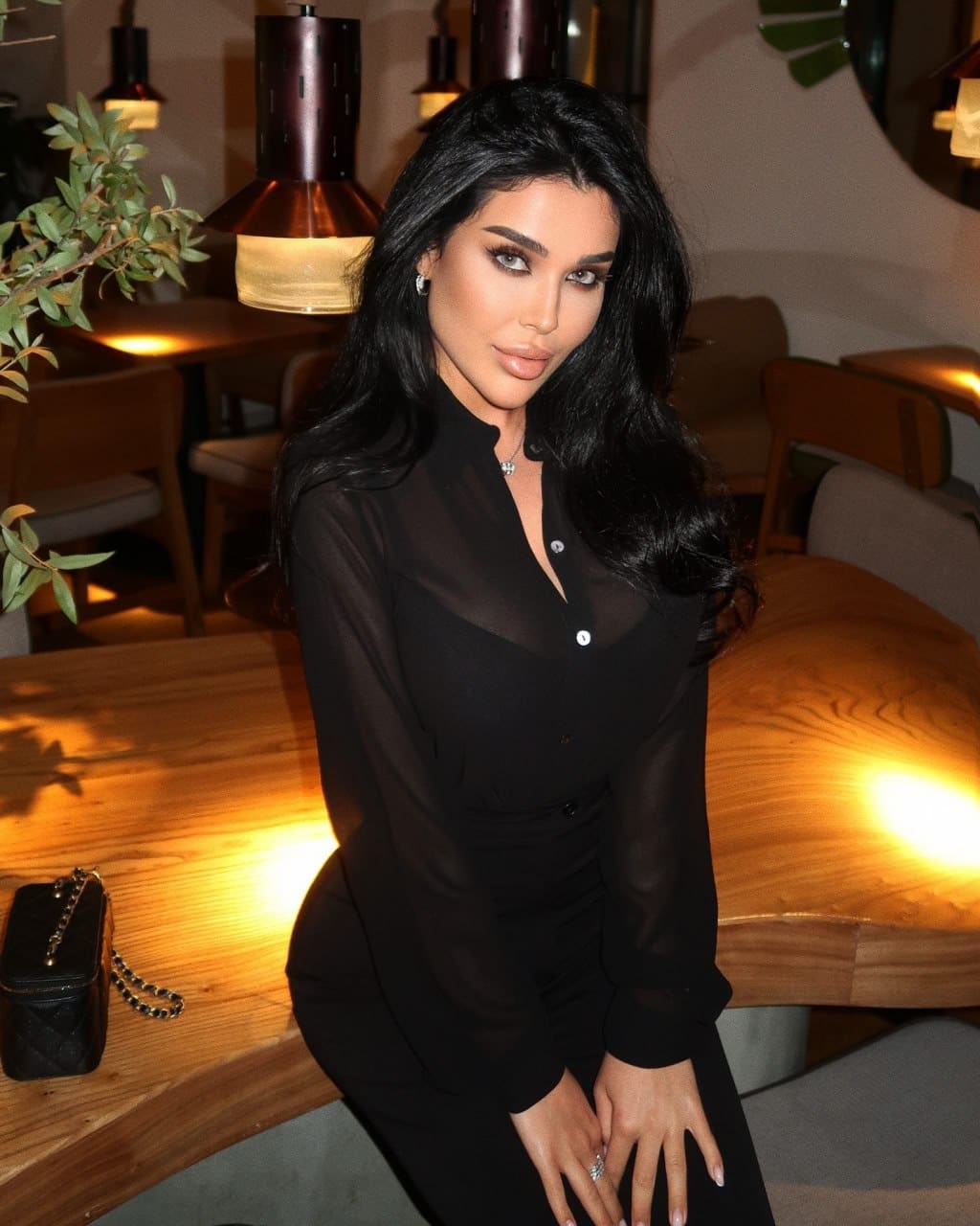 A woman with long dark wavy hair sits at a wooden table in warm light, wearing a sheer black blouse over a black top, with a quilted purse beside her.