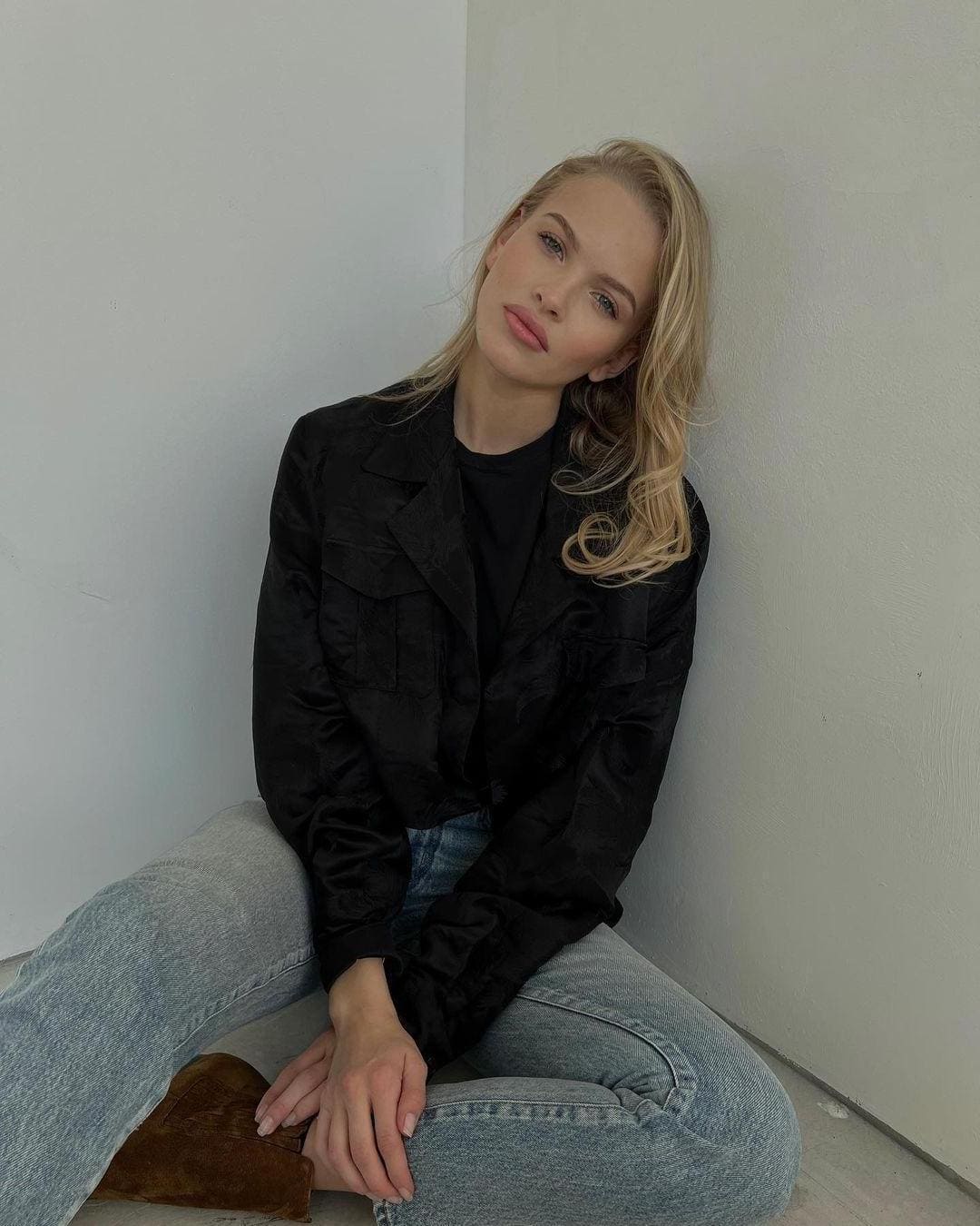 A blonde woman sits on the floor against a white wall, wearing a black jacket, light jeans, and brown suede boots with hands on her lap.