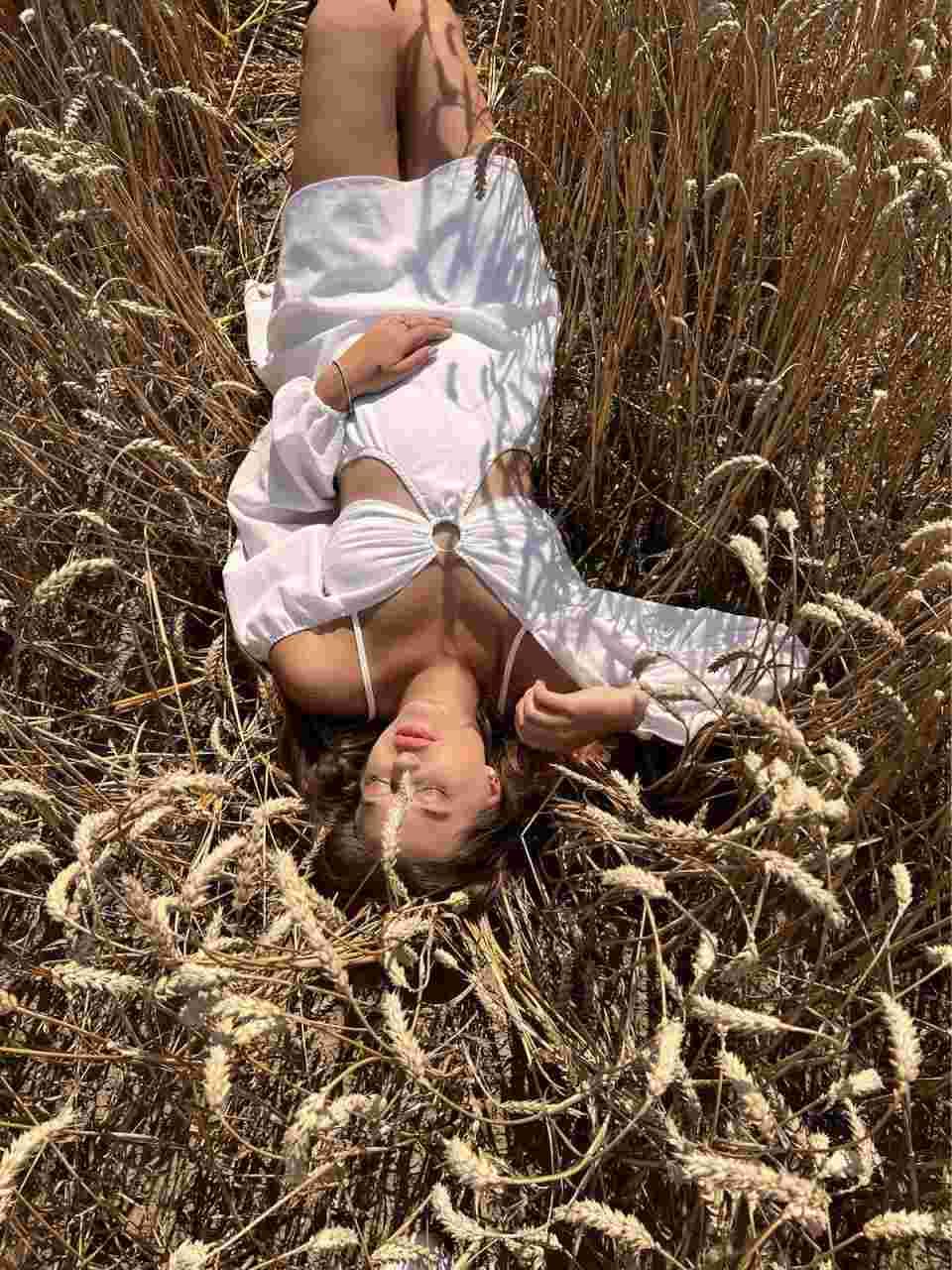 A woman lies on her back in dry wheat wearing an open white long-sleeved top over a white bra, legs bent and one hand on her chest.