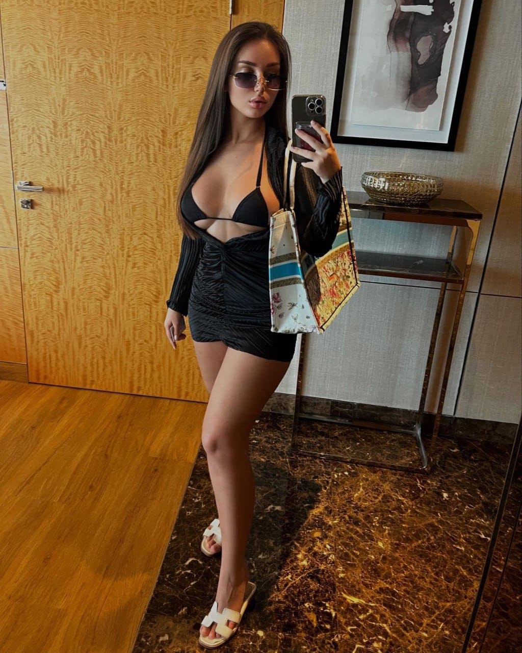 A woman takes a mirror selfie in a black ruched mini dress with a cutout neckline, holding a phone and patterned tote on a marble floor.