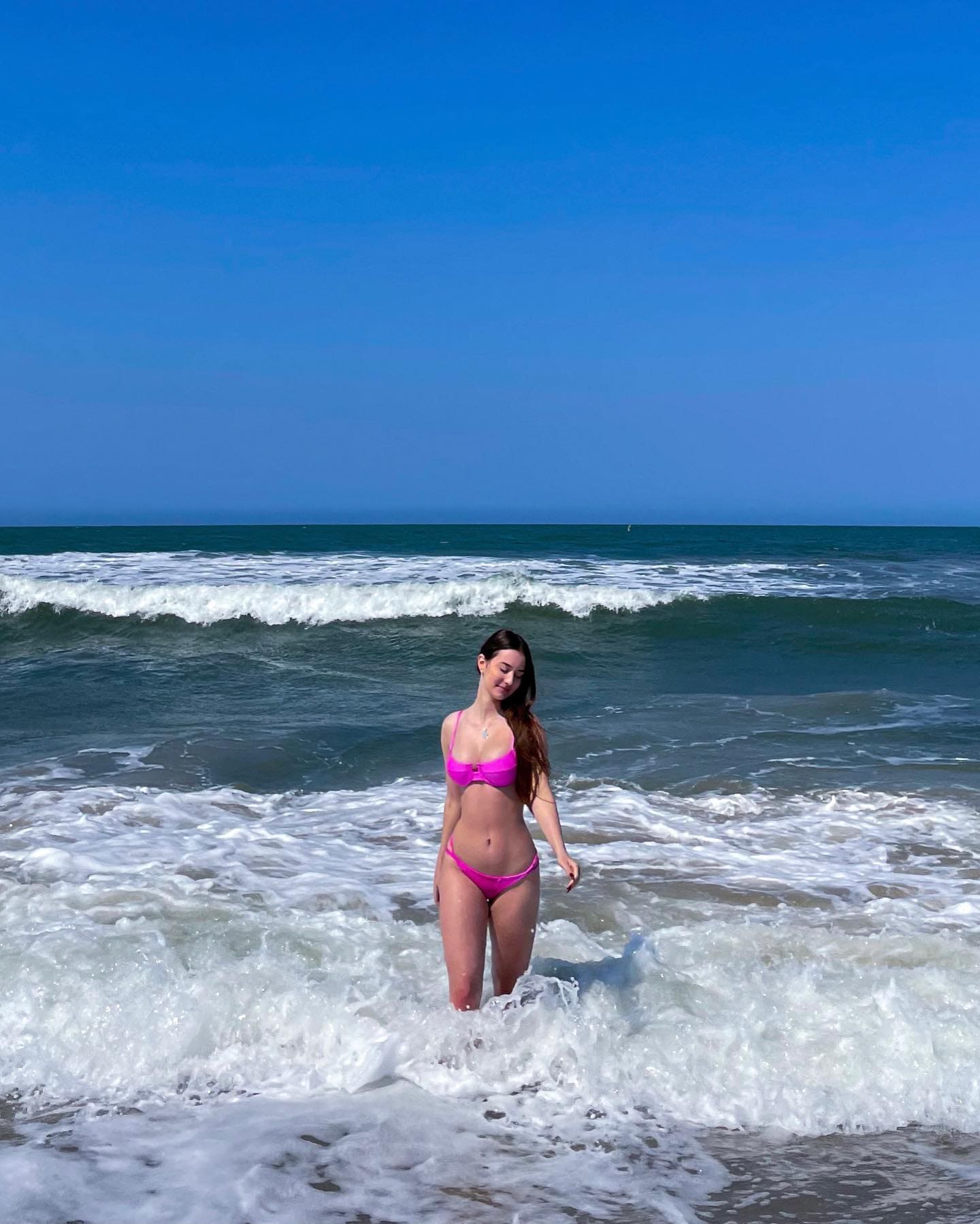 Woman in a bright pink bikini stands in shallow surf, waves breaking around her legs under a clear blue sky.