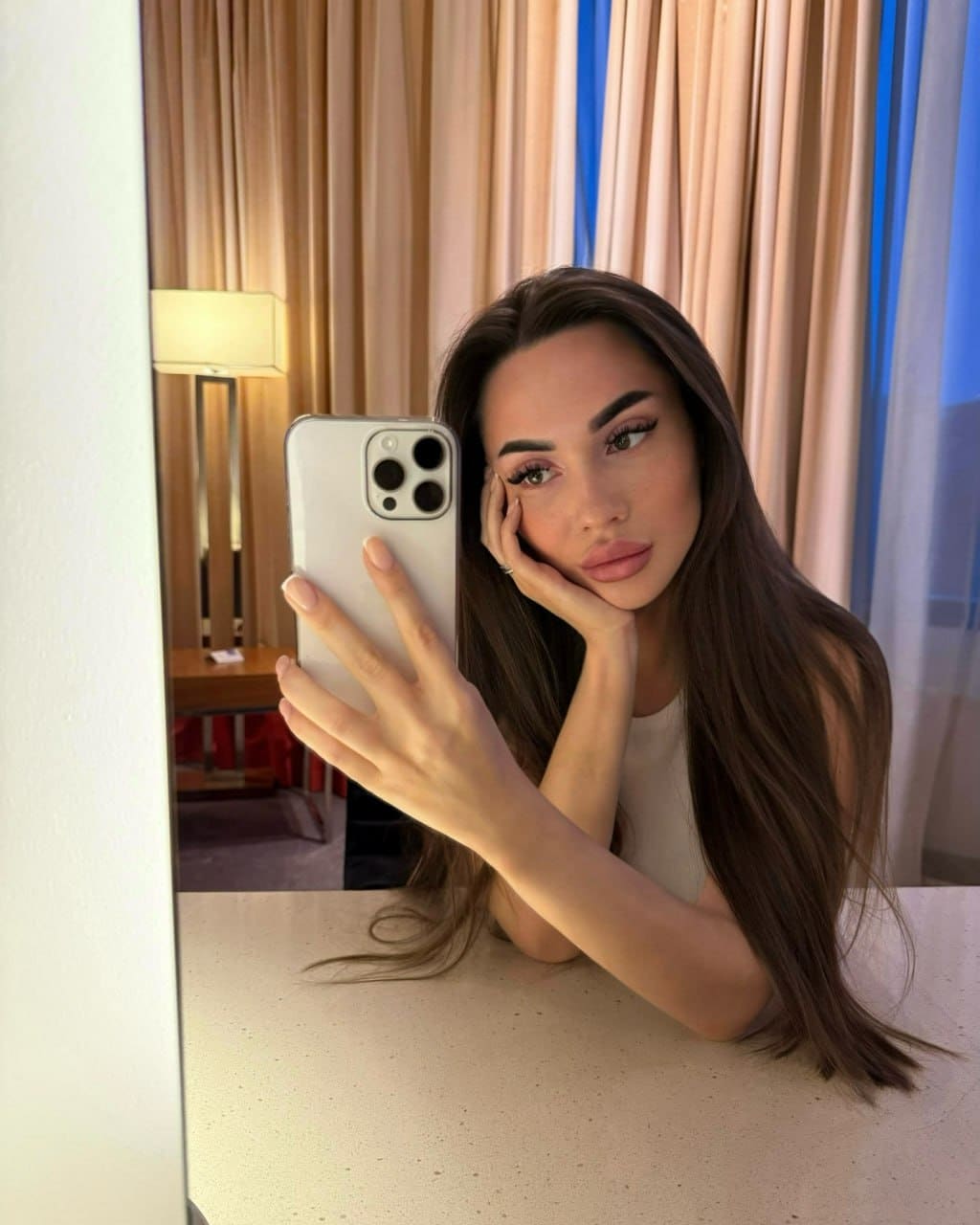 Mirror selfie of a woman resting her chin on her hand, wearing a light sleeveless top against a background of beige curtains and a lamp.