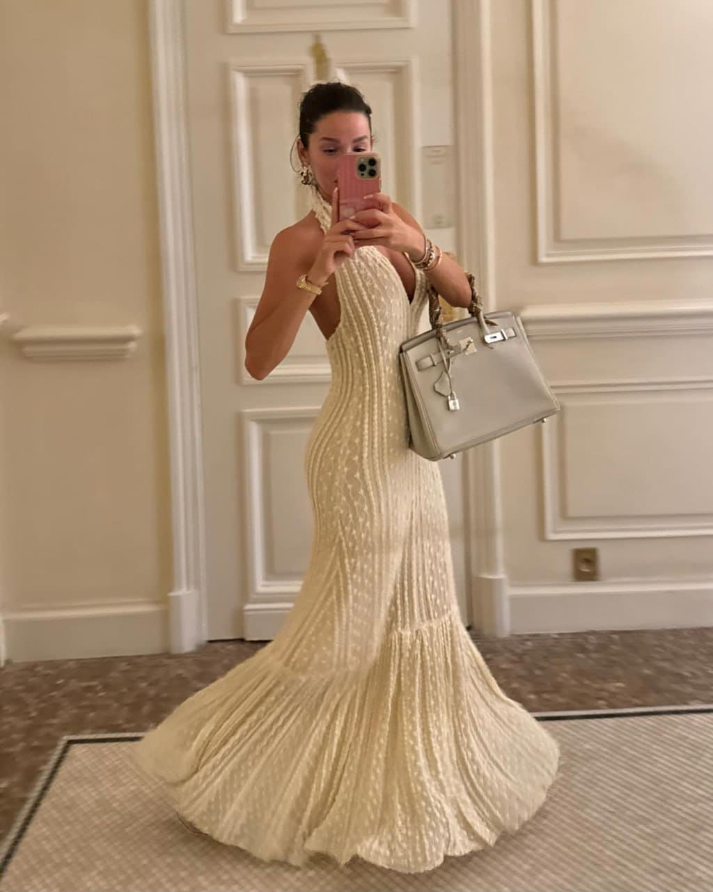 A woman takes a mirror selfie in a cream halter gown, holding a pink phone and gray handbag in a paneled room.
