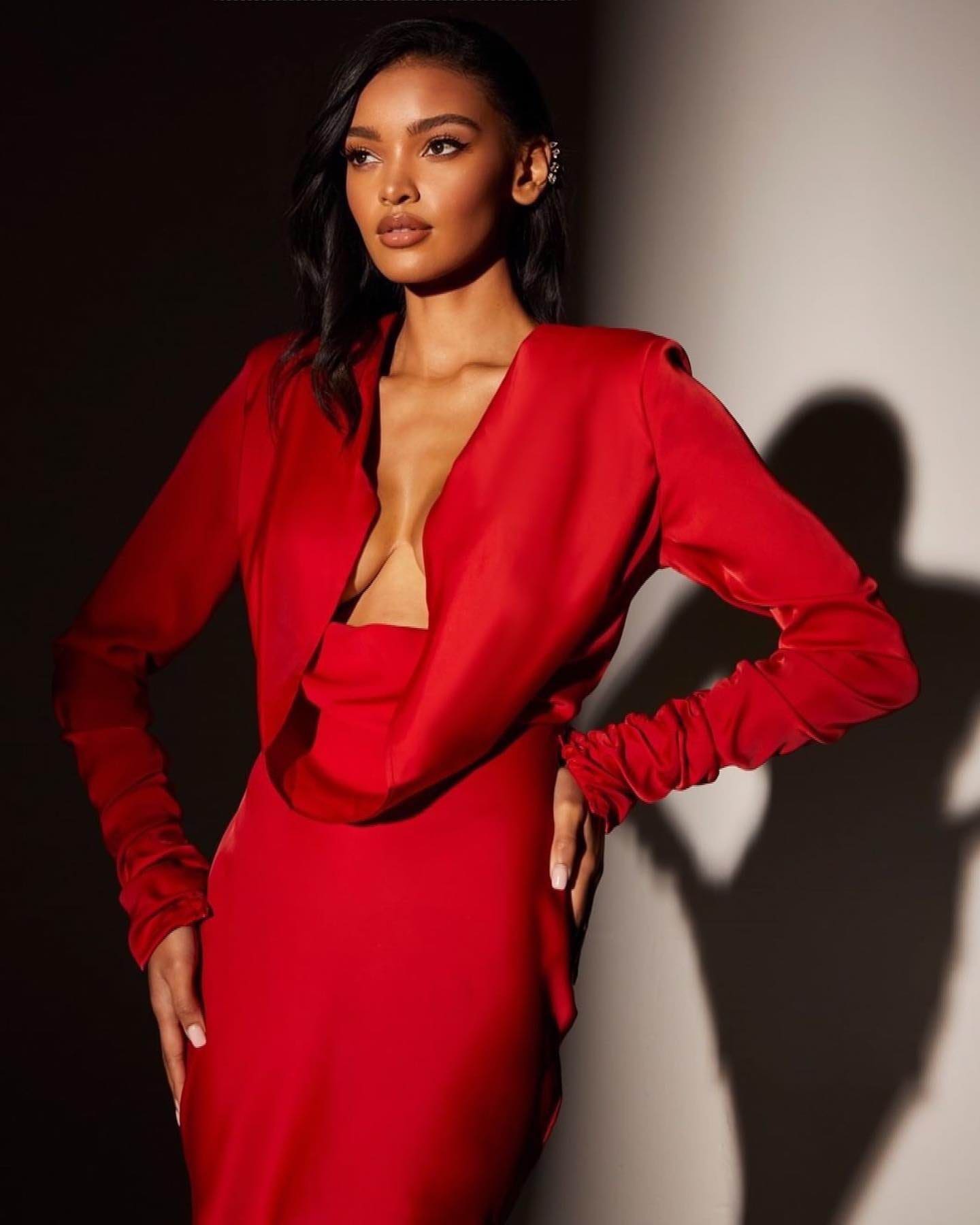 Standing with hands on hips, a woman wears a long-sleeved red dress with a deep V-neck against a plain wall.