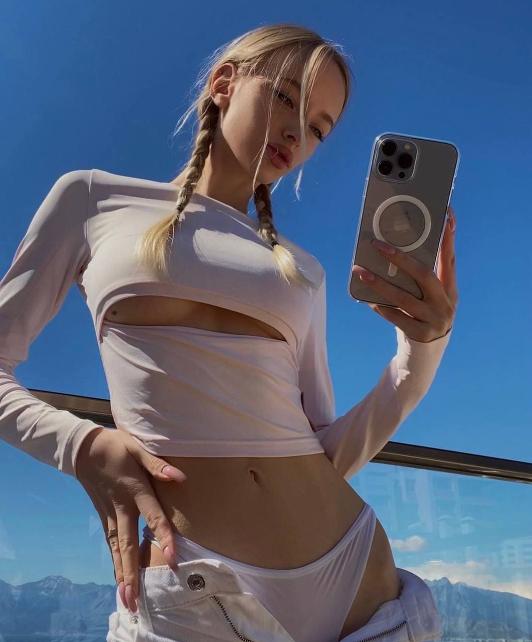 A blonde woman with braids takes a low-angle mirror selfie outdoors against a blue sky, wearing a white crop top with a midriff cutout.