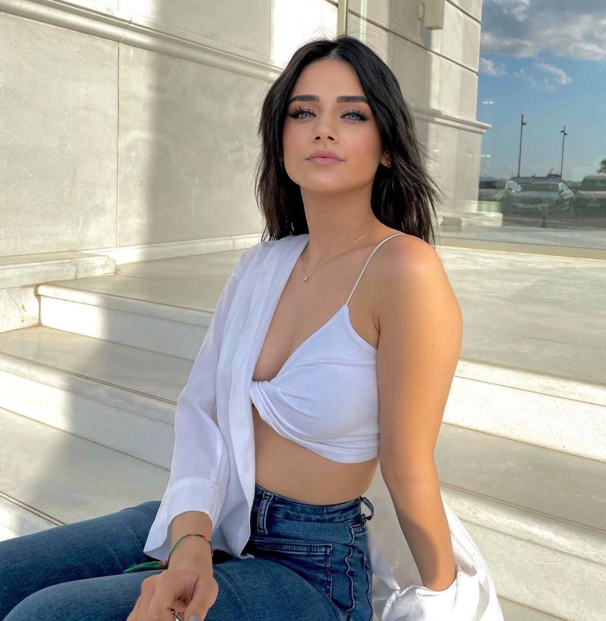 A woman sits on stone steps in a white crop top, open shirt, and jeans, with cars in the background.