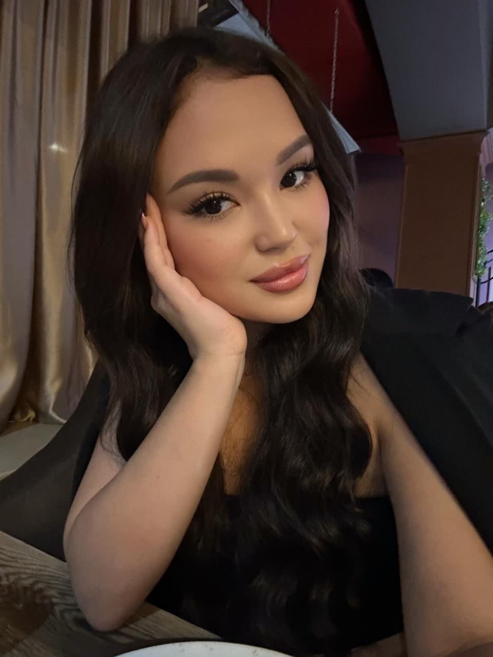 Seated indoors near a curtain, she rests her chin on her hand, wearing a black sleeveless top and a black jacket draped over her shoulders.