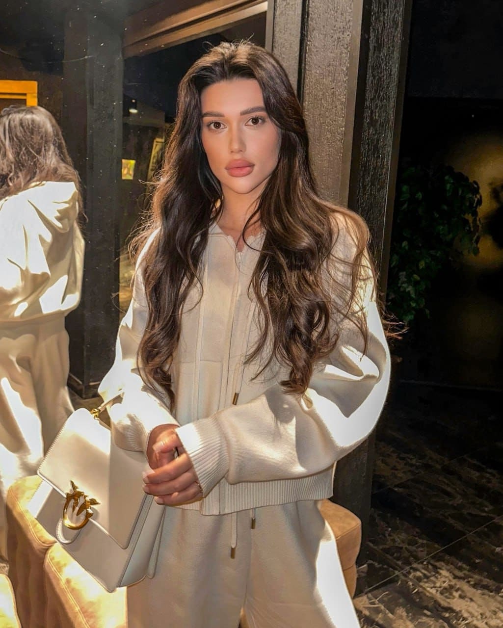 Irina stands facing the camera in a cream zip-up tracksuit, holding a white handbag with a gold clasp against a dark interior background.