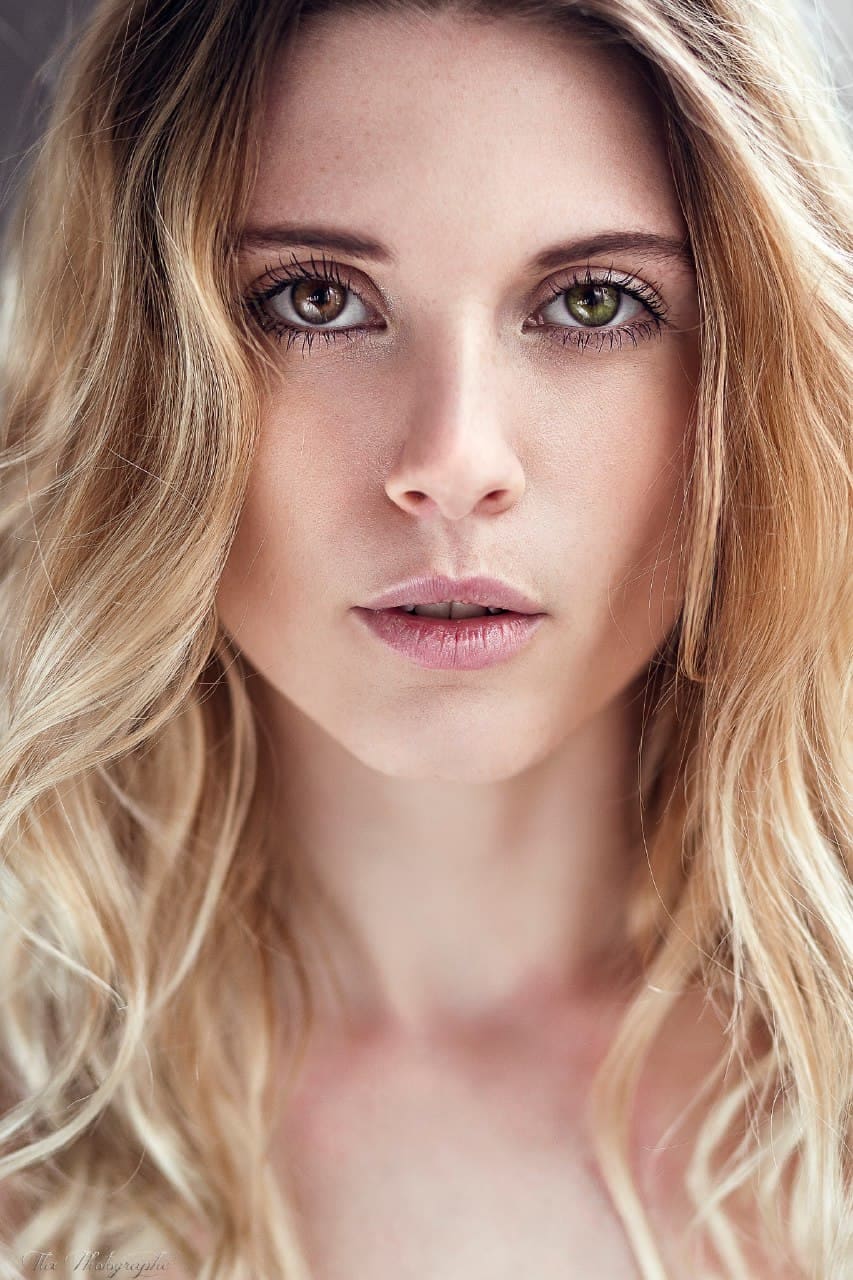 Close-up portrait of a woman with wavy blonde hair and heterochromia, looking at the camera with bare shoulders against a blurred background.