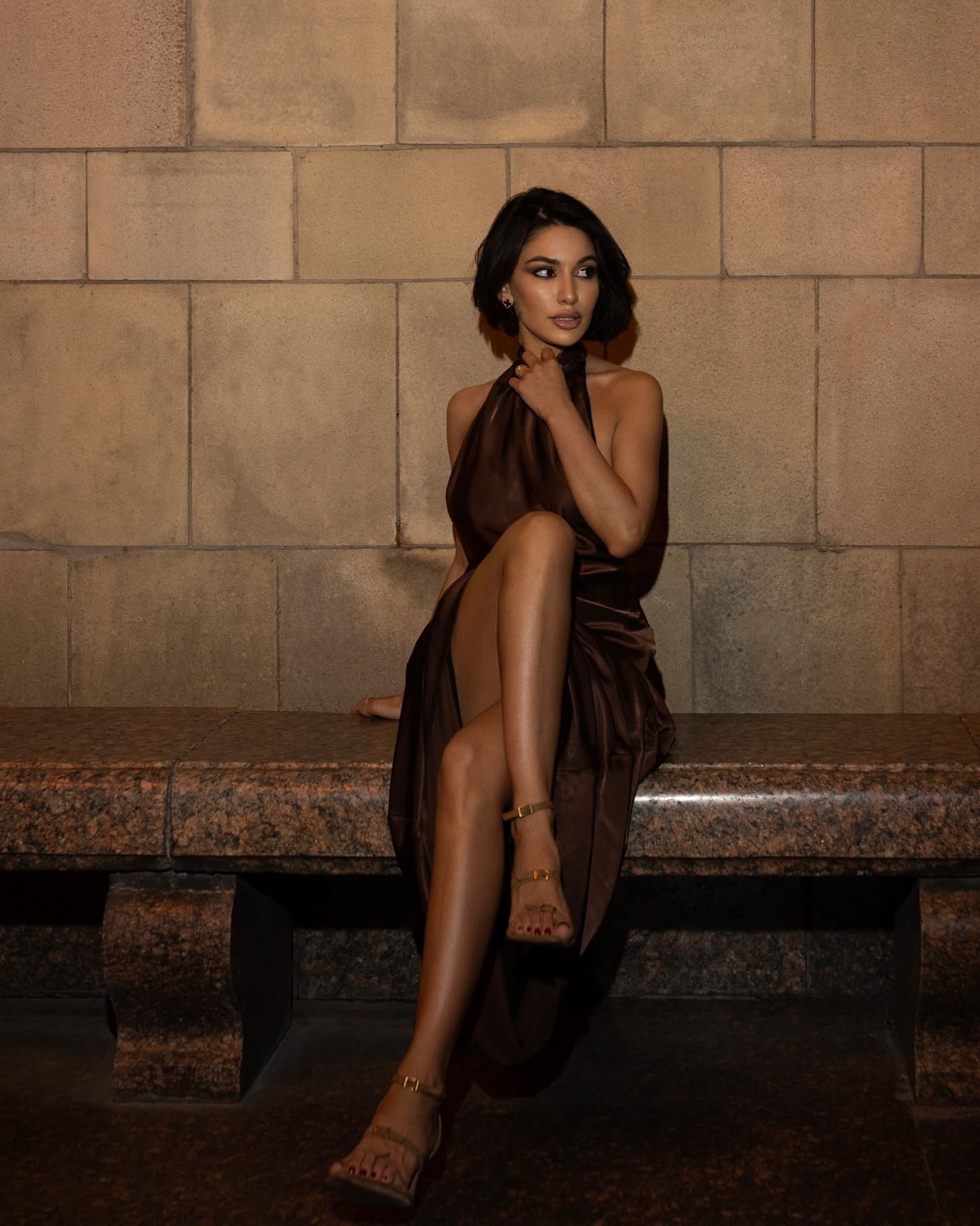 Seated on a stone bench against a stone wall, she wears a dark brown halter-neck dress with a high slit and strappy sandals.