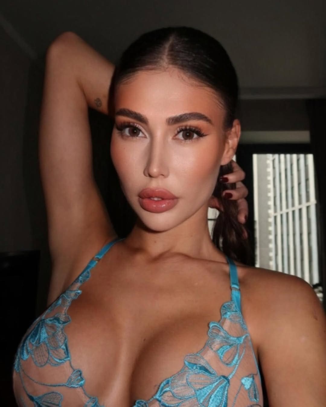 Seraphina poses with one arm raised behind her head, wearing a blue lace halter top in a dimly lit room with blinds.