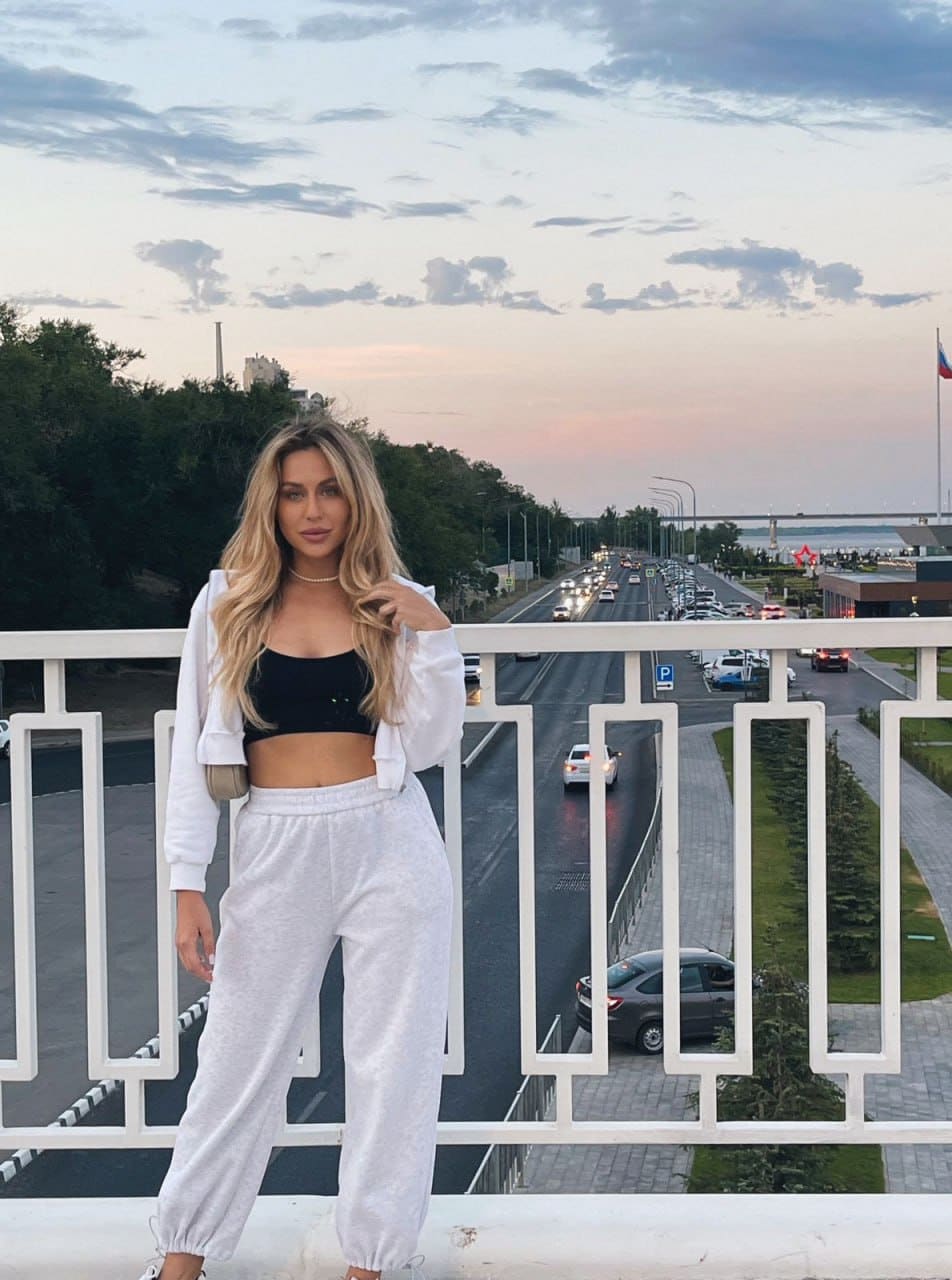 A woman stands on a bridge railing in a black crop top, white sweatpants, and open hoodie, with a highway and cloudy sunset sky in the background.