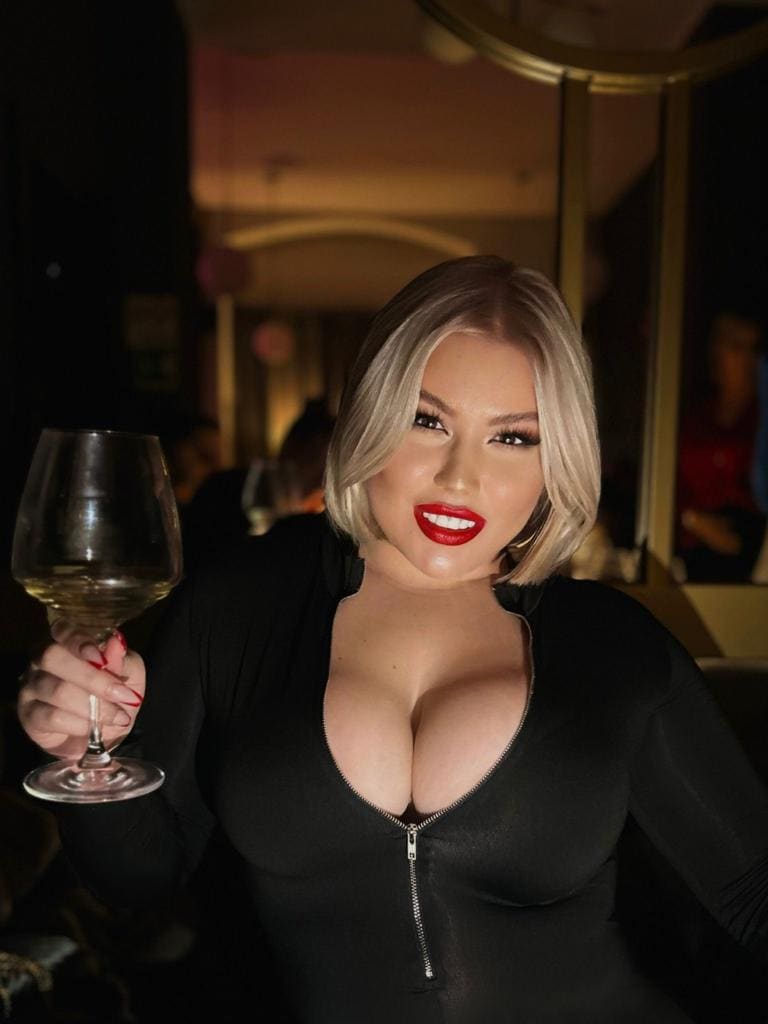 A woman with a blonde bob and red lipstick holds a wine glass, wearing a black zippered top with a plunging neckline in dim light.