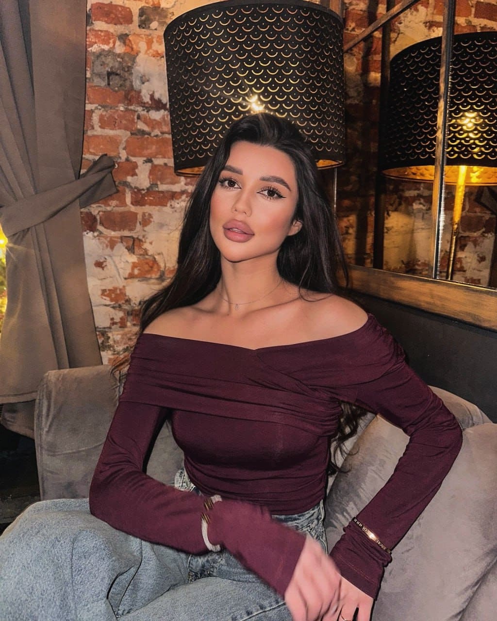 A woman sits on a grey couch wearing a maroon off-the-shoulder top and light blue jeans, with a brick wall and patterned lamps behind her.