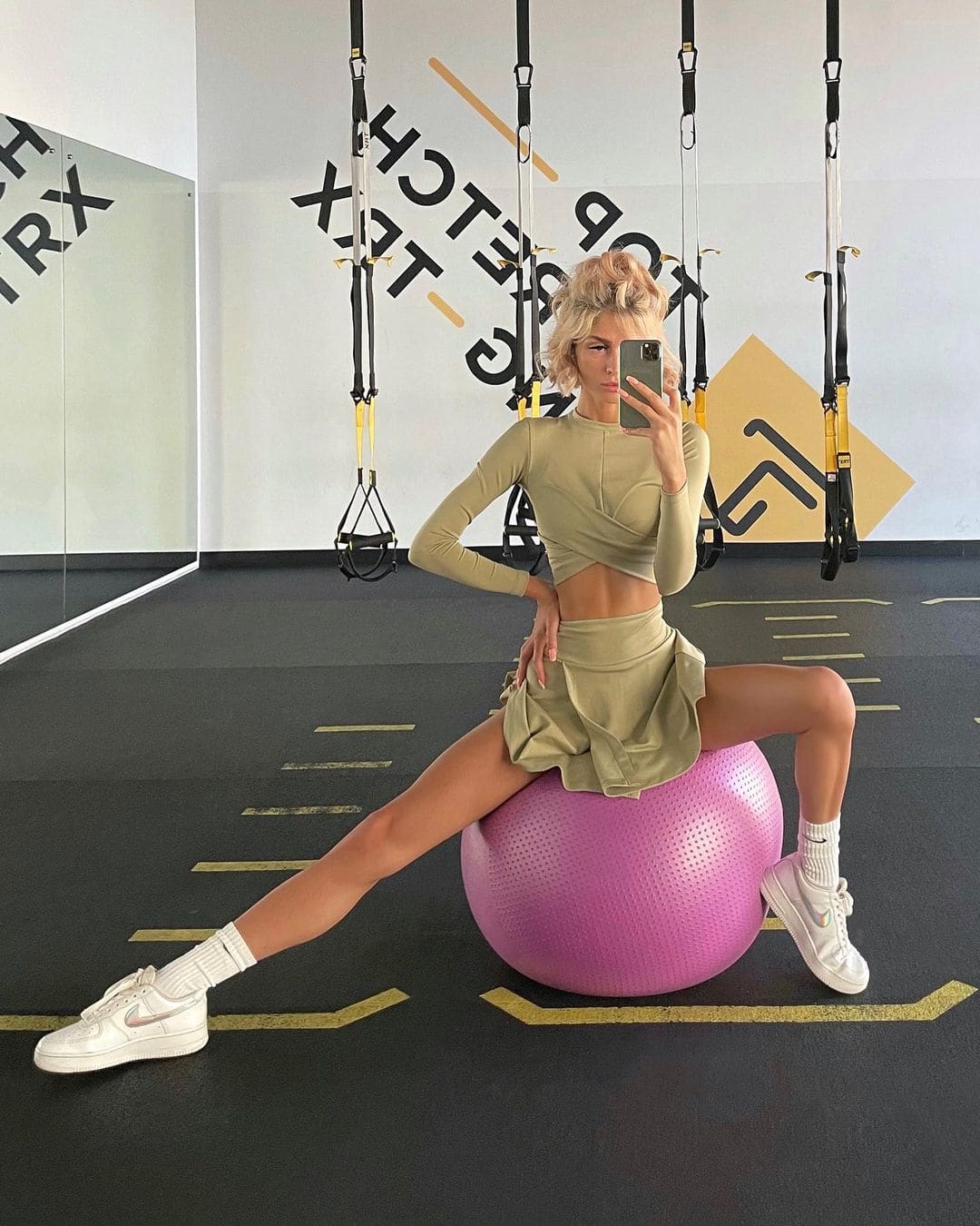 A blonde woman sits on a pink exercise ball in a gym, wearing a cropped top and skirt while taking a mirror selfie.