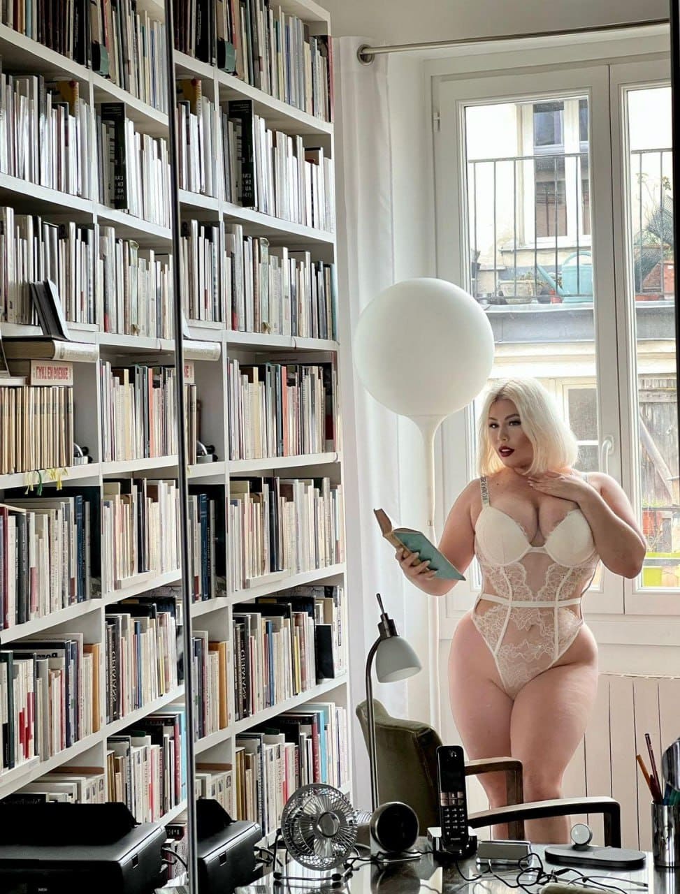 A blonde woman in a white lace bodysuit stands by bookshelves, holding an open book and touching her neck, with a desk behind.