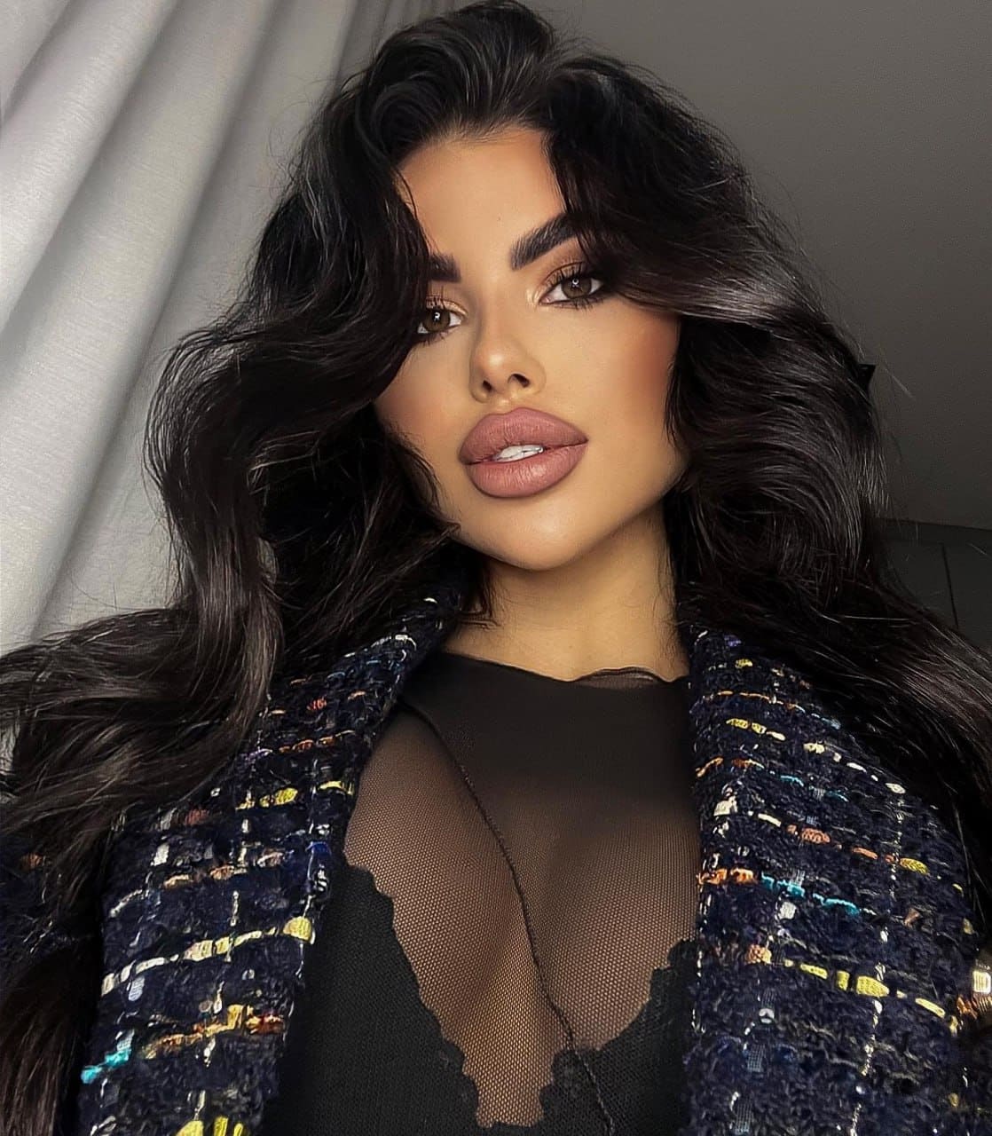 A woman faces the camera in a tweed jacket over a sheer black top, revealing a lace bra, against light curtains and a gray wall.