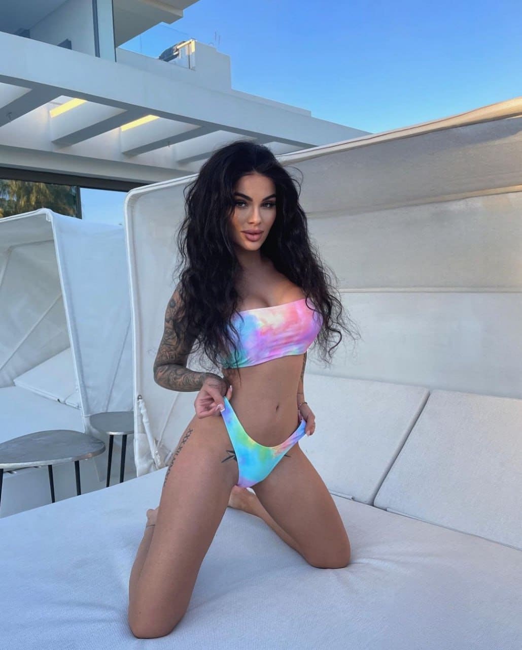 A woman kneels on a white outdoor lounge in a pastel tie-dye bikini, with a white building and blue sky behind.