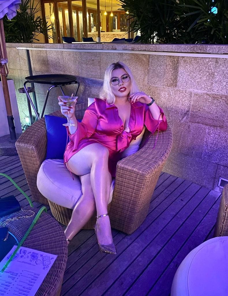 A blonde woman in glasses and a pink satin dress sits in a wicker chair on a deck, holding a cocktail with one hand near her face.