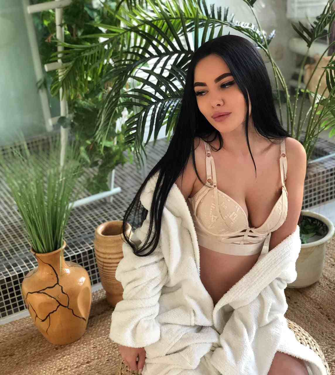 A woman sits on a woven stool wearing a beige strappy bra and open white bathrobe, with potted plants and a tiled wall visible behind her.