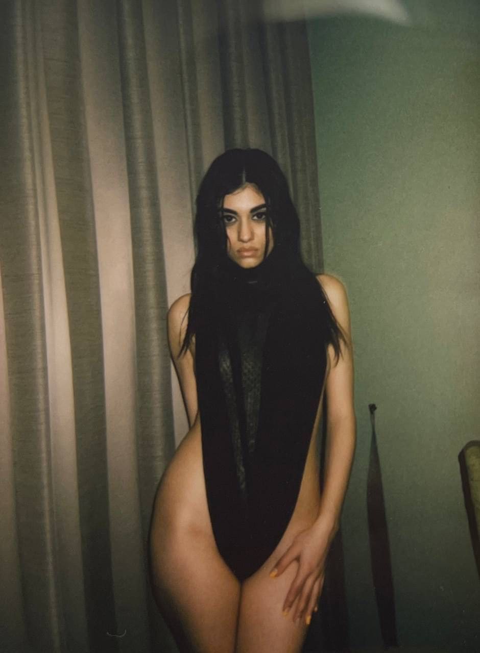 Standing in a dim room with a curtain behind her, she wears a black sleeveless top with a sheer front panel, hand on thigh.