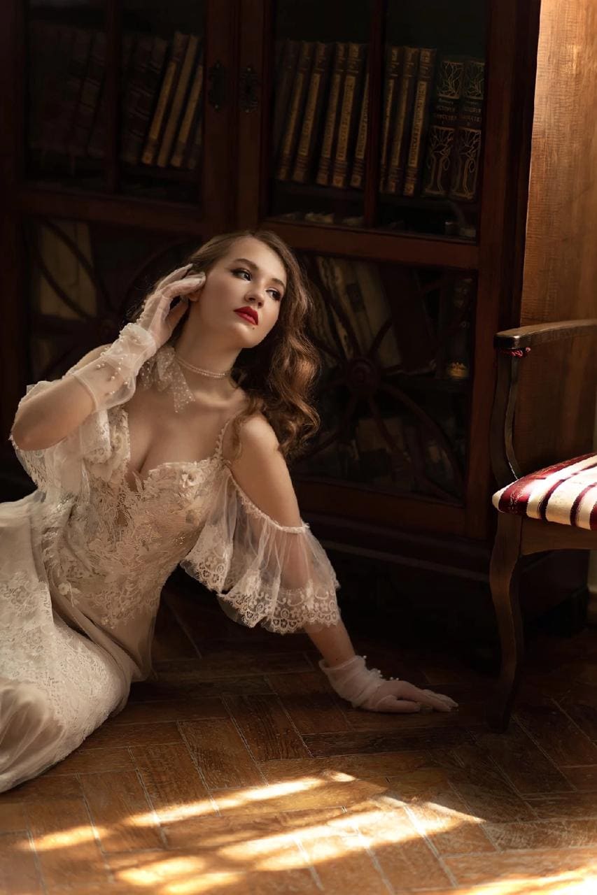 A woman sits on a wooden floor before a bookcase in a sheer white lace dress and gloves, one hand touching her hair.