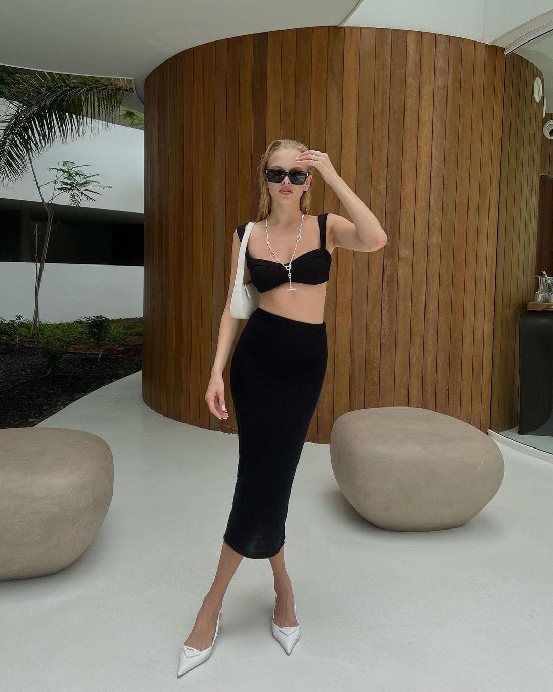 A blonde woman in sunglasses stands before a curved wooden wall, wearing a black bustier, midi skirt, and white heels with one hand raised.