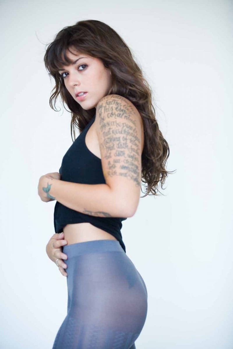 Nova stands in profile against a white background, wearing a black cropped tank top and sheer grey leggings, looking over her shoulder.