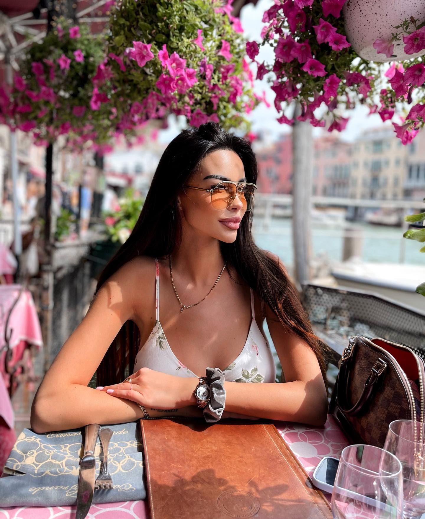 Seated at an outdoor table near water, she wears a white floral top and aviator sunglasses, with pink flowers above.