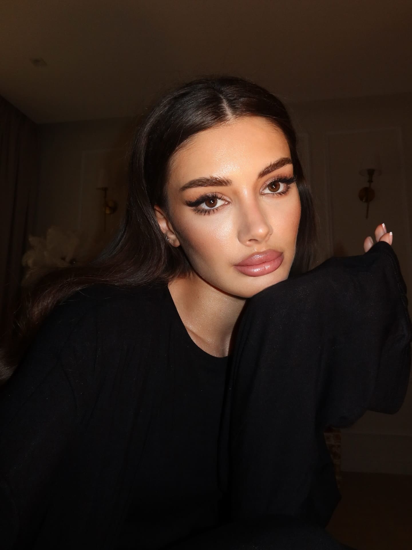 A woman with long dark hair rests her chin on her hand while wearing a loose black top in a dimly lit room with wall sconces.