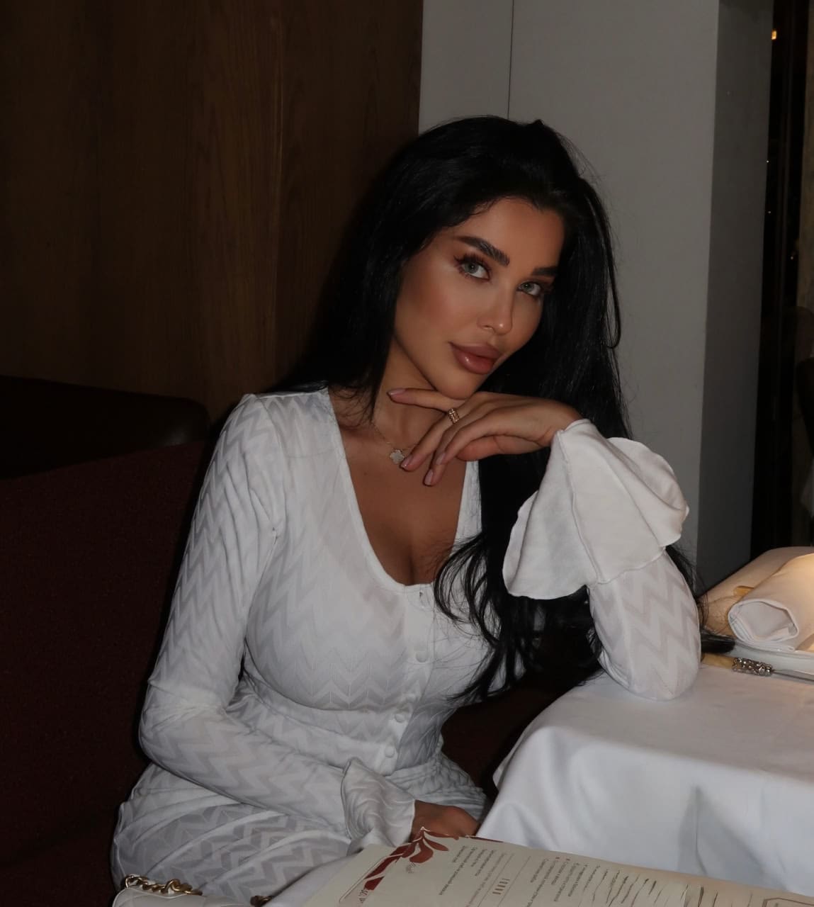 A woman sits at a table in a dim room, wearing a white long-sleeved dress with a chevron pattern and deep V-neck, resting her chin on her hand.