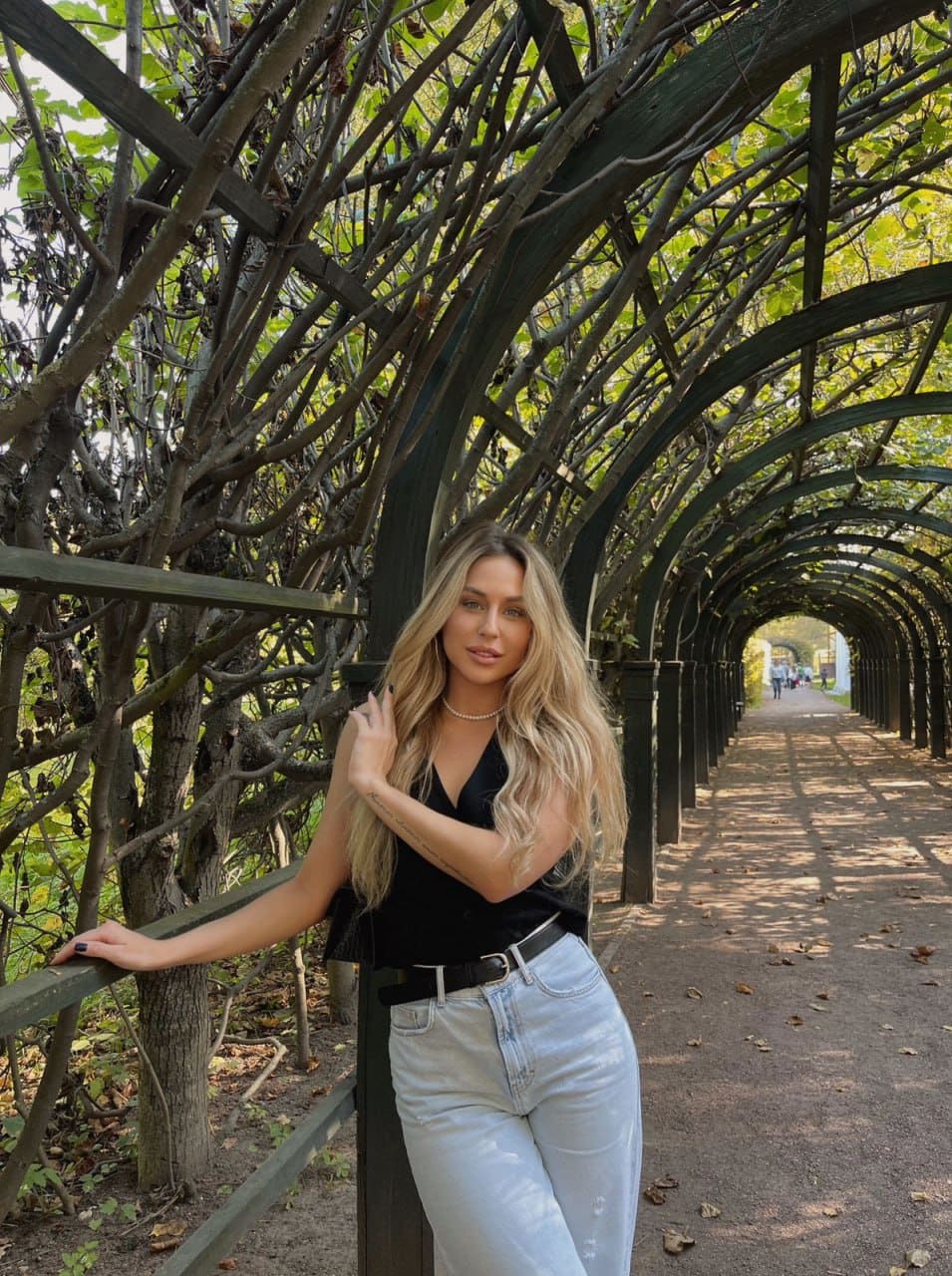 A woman stands in a leafy garden archway wearing a black sleeveless top and light jeans, resting one hand on the wooden railing.