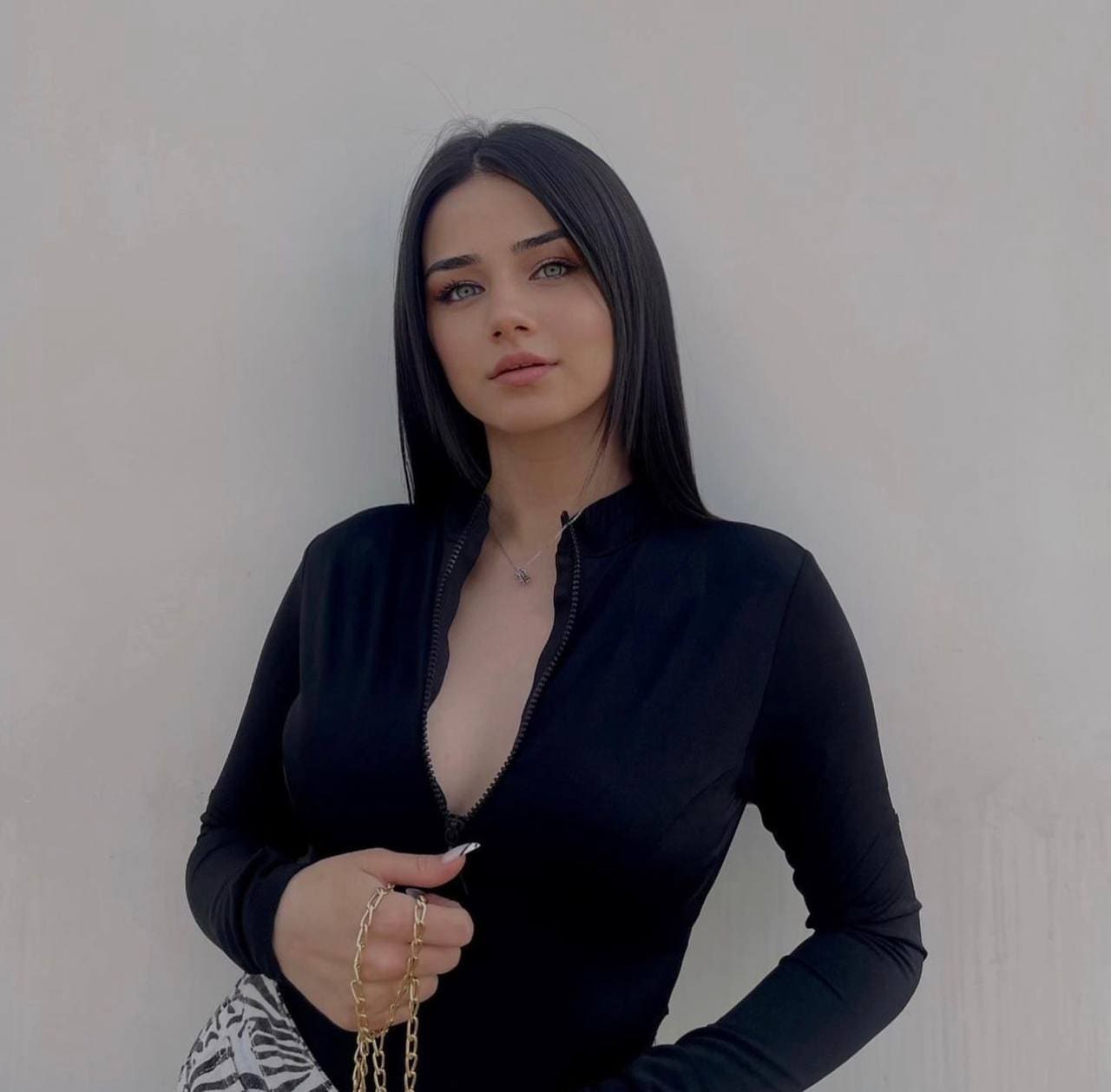 A woman in a partially unzipped black zip-up top holds a gold chain strap against a plain wall.