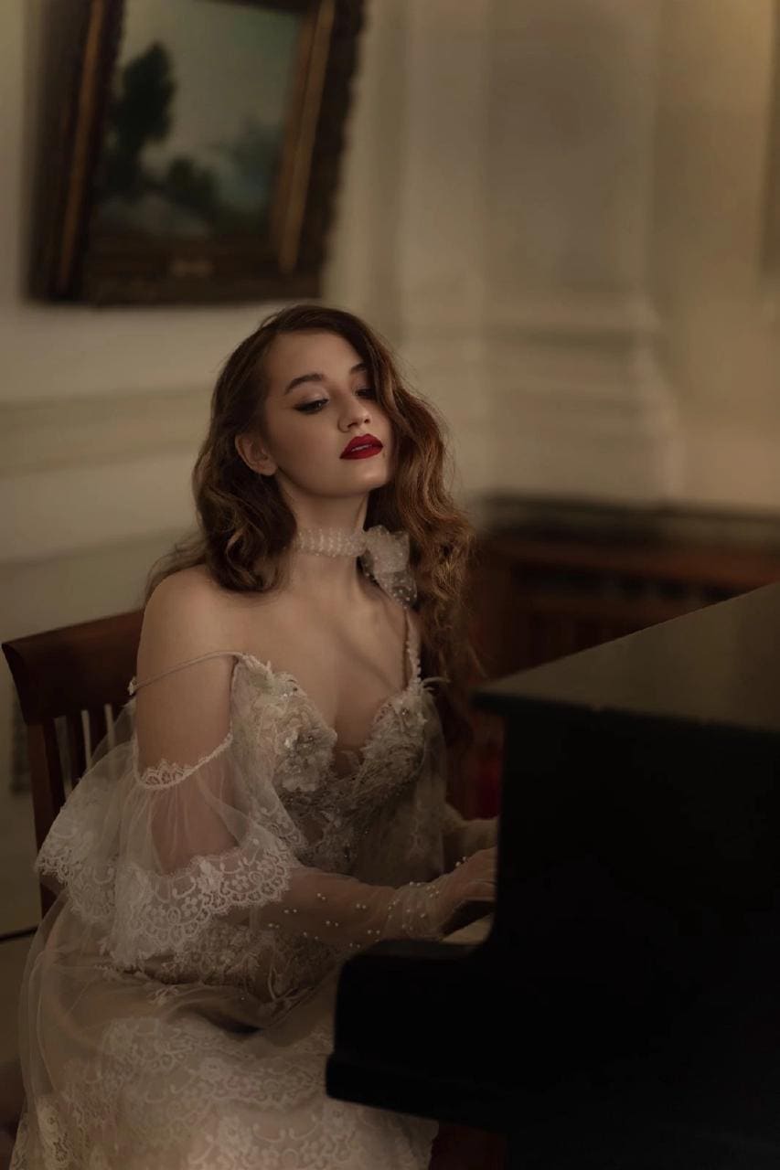 A woman in a white lace off-shoulder dress sits at a dark piano, with a framed painting on the wall behind her.