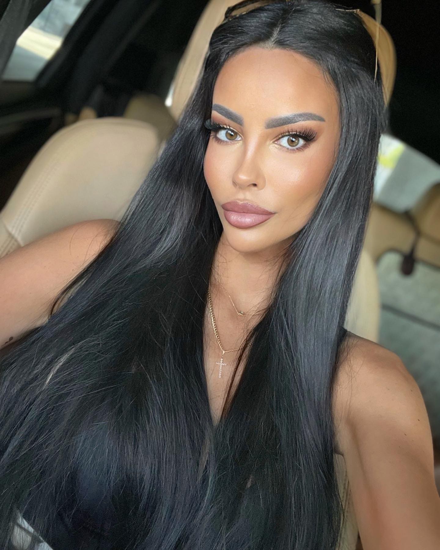 Cheyenne sits in a car's beige leather back seat, wearing a dark off-shoulder top and a gold cross necklace.