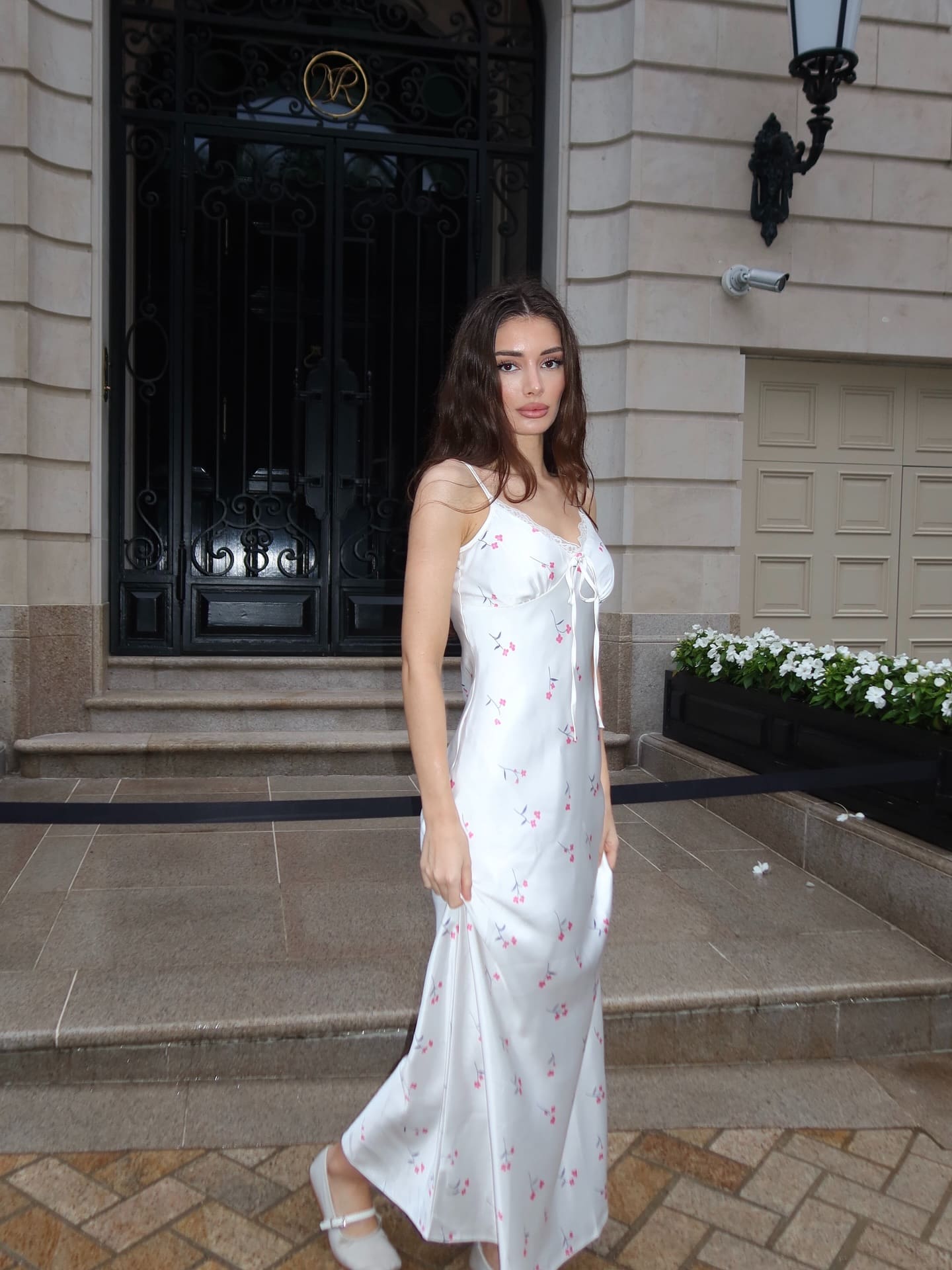 Sabina stands on a stone step before a black iron gate, wearing a white sleeveless floral dress and white shoes.