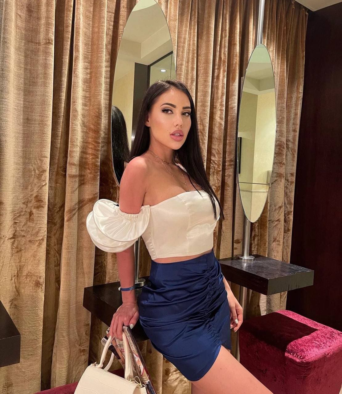 Nikitta stands in a room with gold curtains and oval mirrors, wearing a white off-the-shoulder crop top, navy skirt, and holding a white handbag.