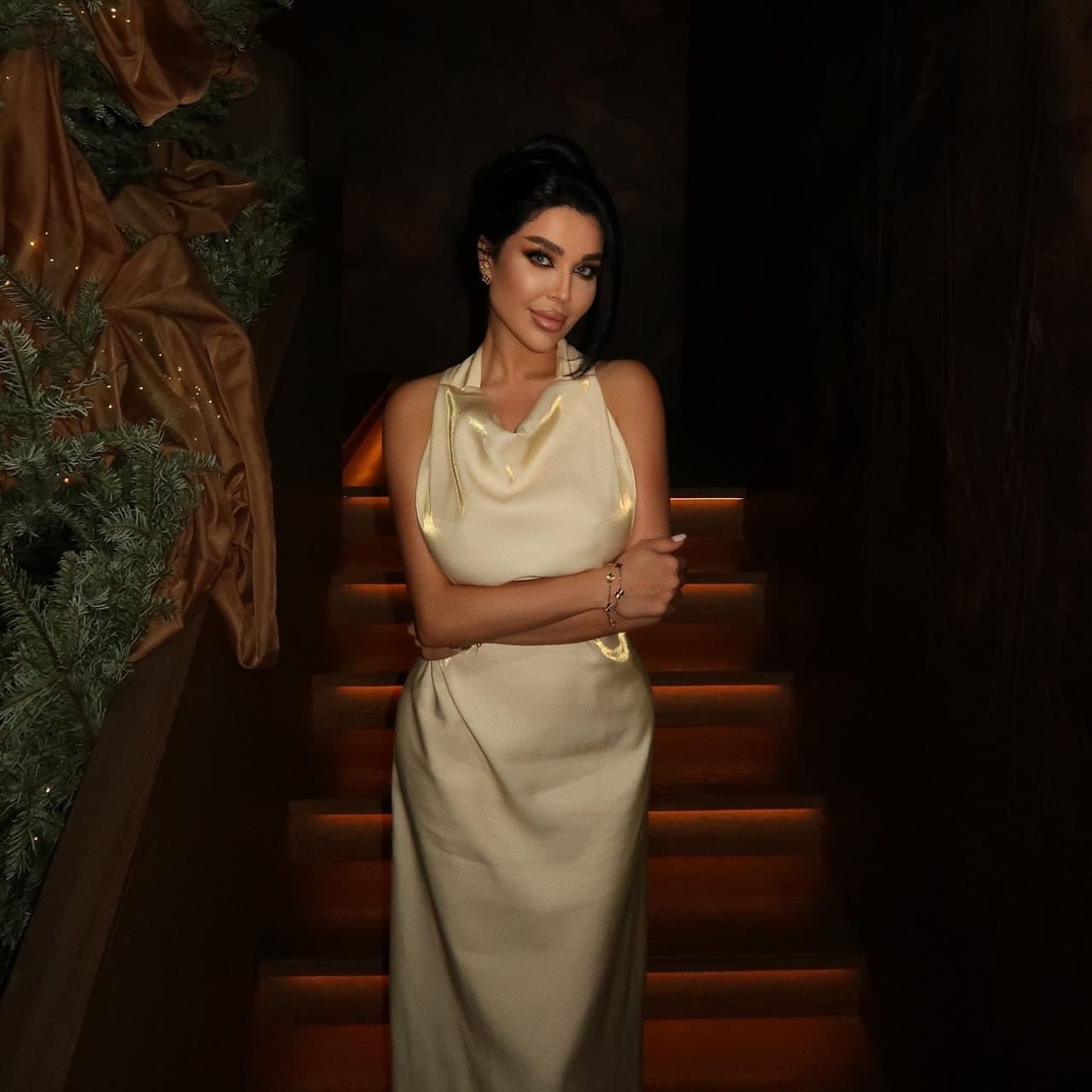A woman with a high bun stands with arms crossed in a sleeveless cream satin halter dress, positioned near a dimly lit staircase and evergreen branch.