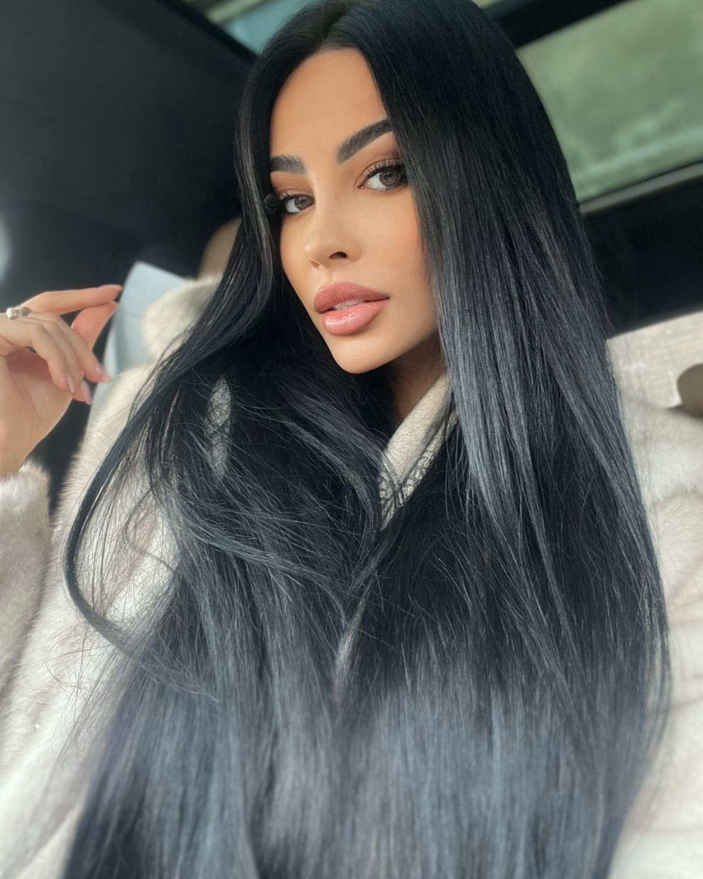 Inside a car, she wears a light fuzzy coat and raises one hand near her face, with straight black hair.