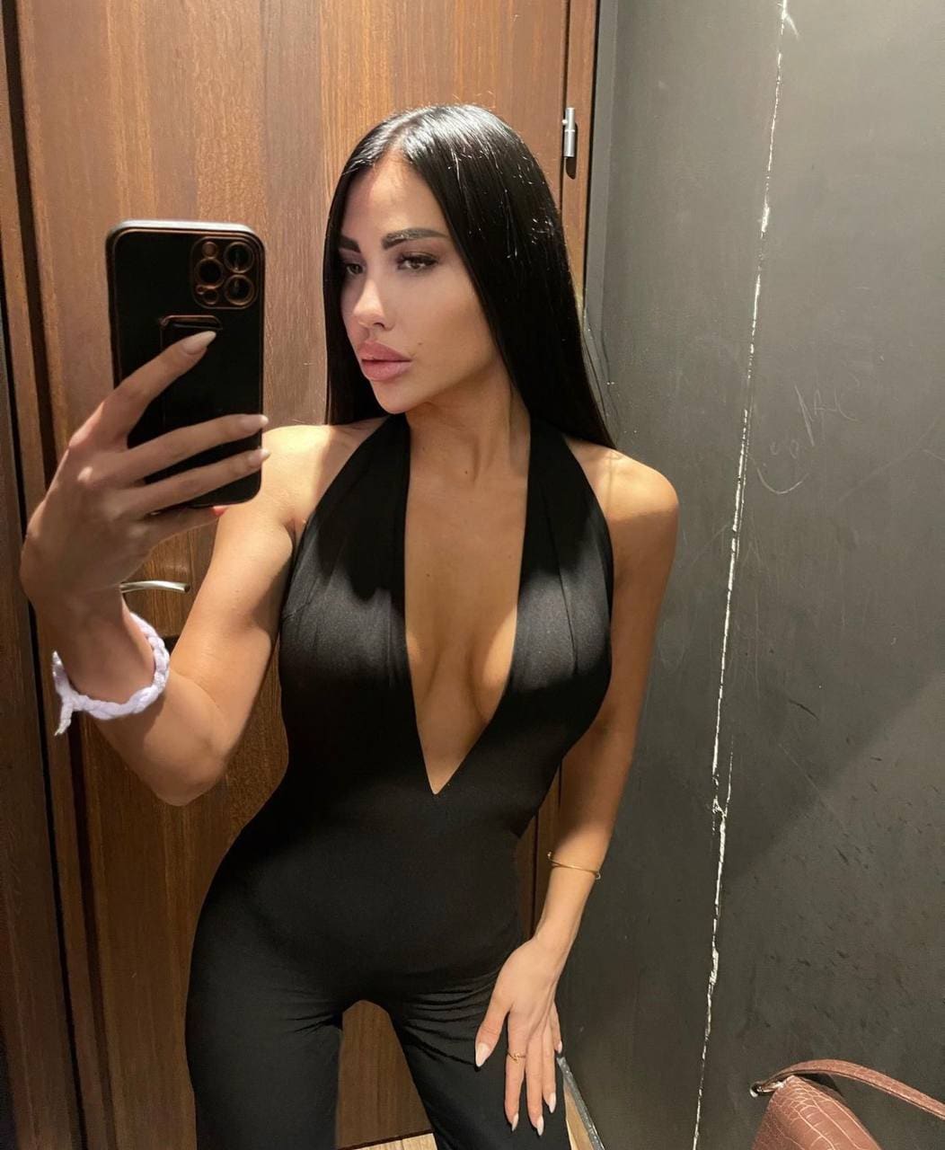 A mirror selfie features a woman in a black halter-neck bodysuit with a deep neckline, standing before a wooden door and dark wall.
