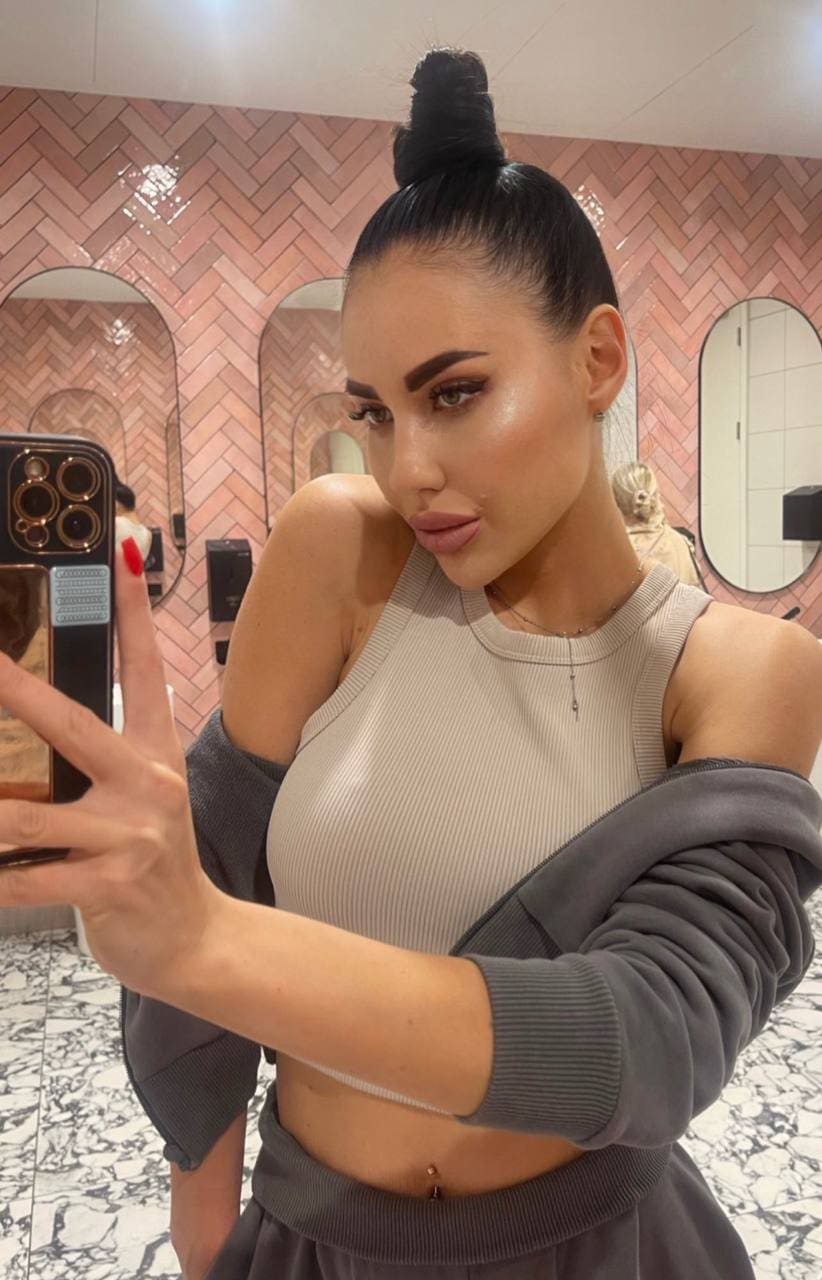 A mirror selfie shows a woman with a high bun in a beige ribbed crop top and grey sweatshirt draped off her shoulders, revealing a belly button ring.